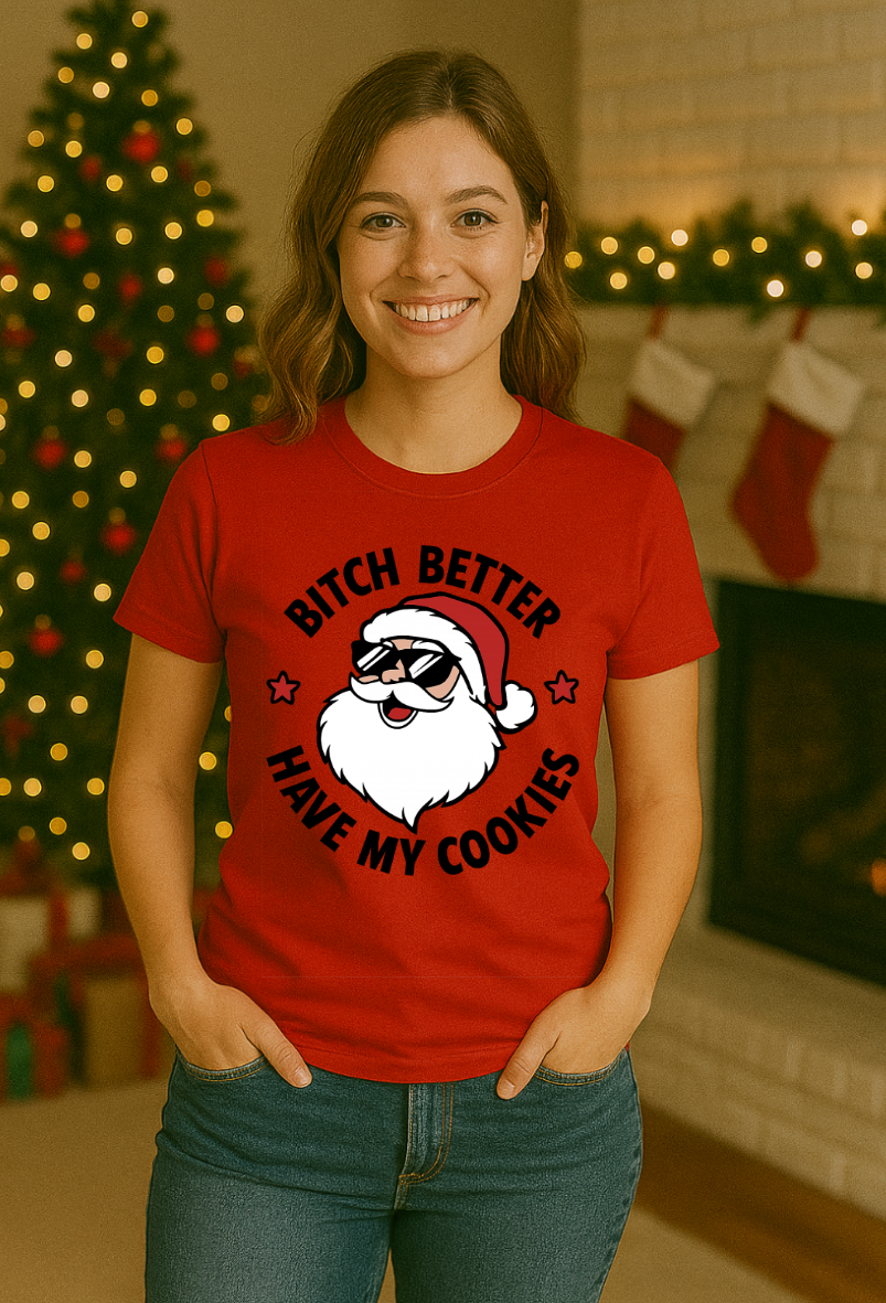 Woman wearing a red Christmas t-shirt featuring a cartoon Santa with sunglasses and a humorous cookie-themed holiday design, standing in a festive indoor setting with a decorated tree and stockings.