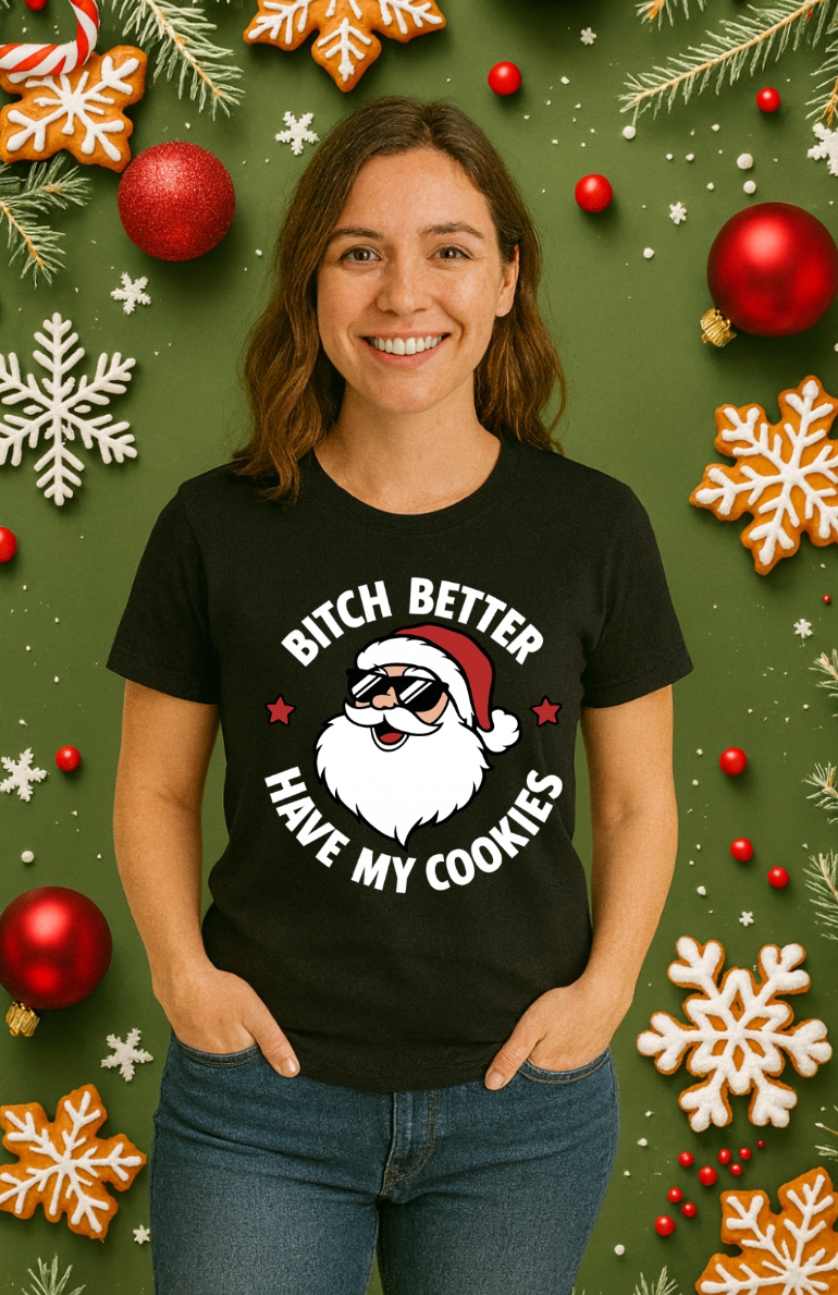Woman wearing a black Christmas t-shirt featuring a cartoon Santa with sunglasses and a humorous cookie-themed holiday design, standing in front of a festive green background with ornaments and snowflakes.