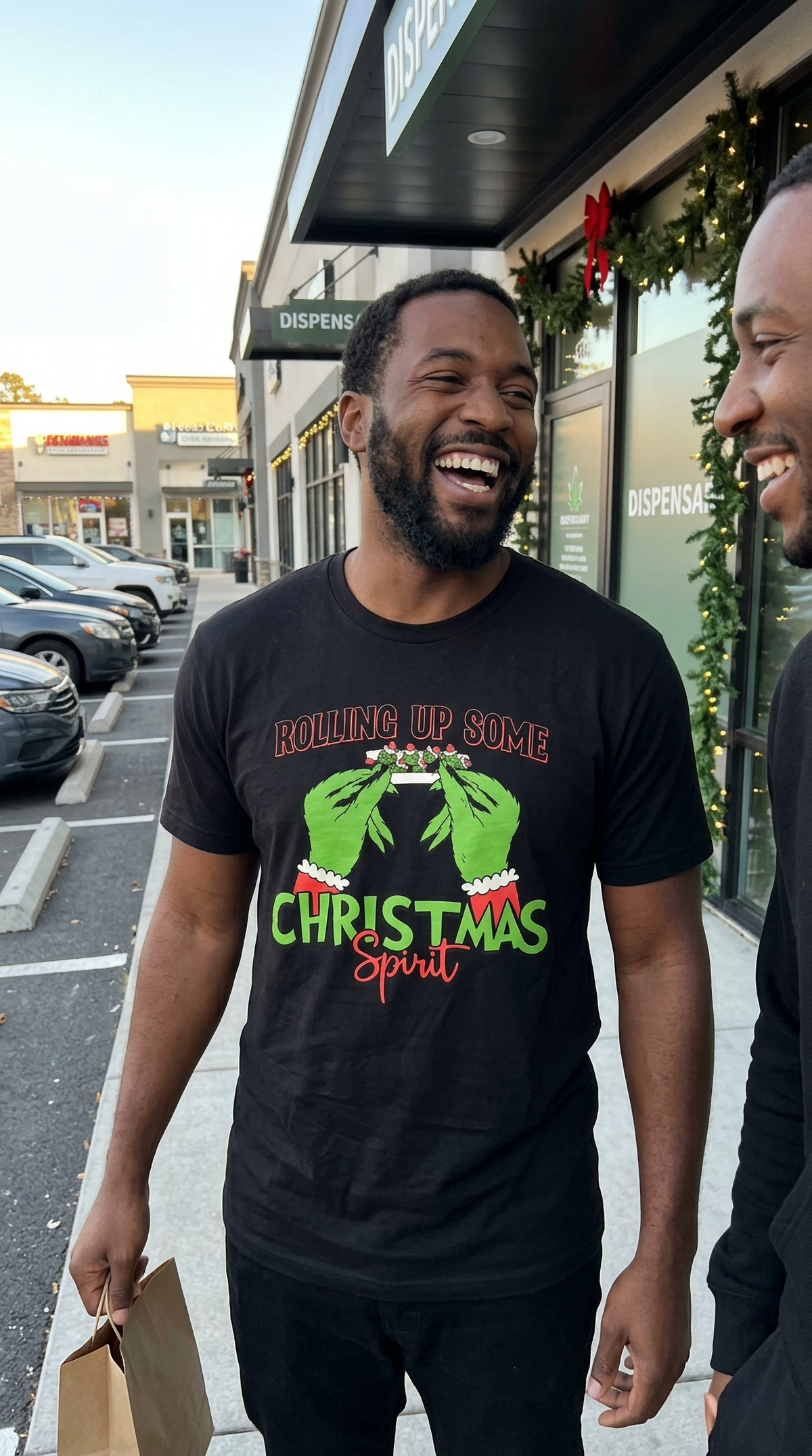 Man wearing black Rolling Up Some Christmas Spirit graphic t-shirt laughing with a friend outside a shopping center.