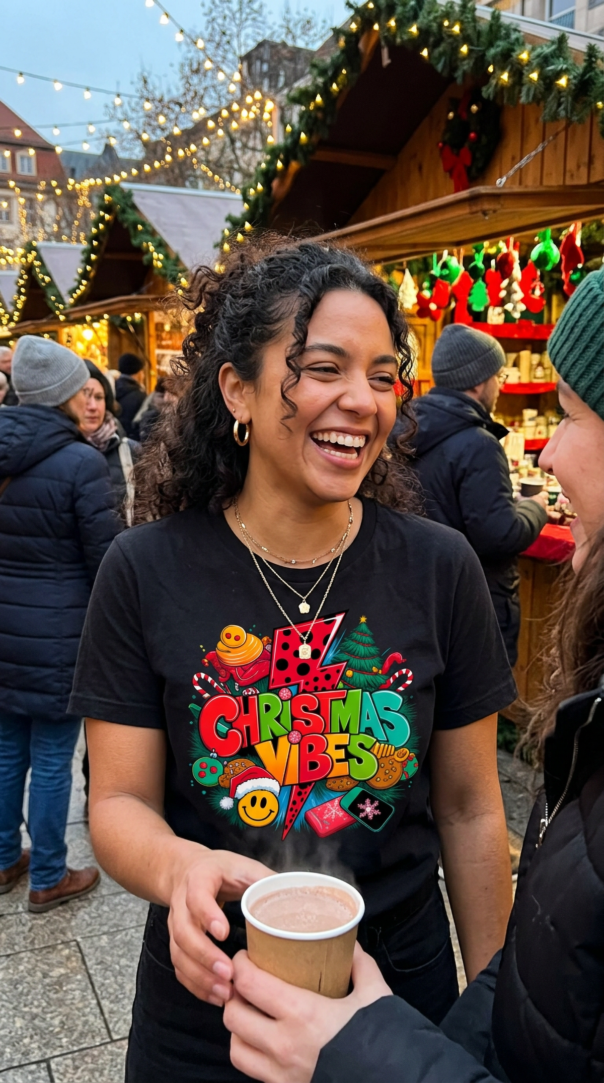 Black: Woman wearing a black Christmas Vibes graphic t-shirt at a festive outdoor holiday market, featuring colorful retro Christmas artwork.