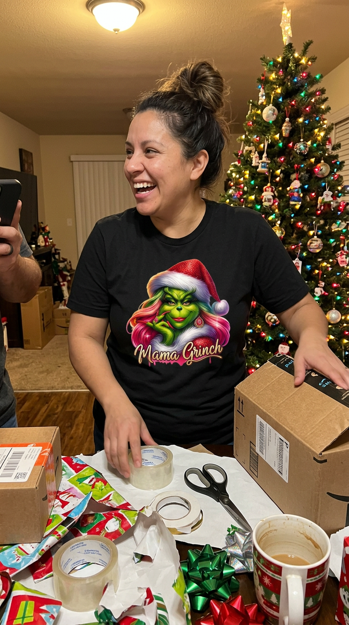 Smiling woman wearing black Mama Grinch Christmas t-shirt while wrapping holiday gifts beside a decorated Christmas tree.