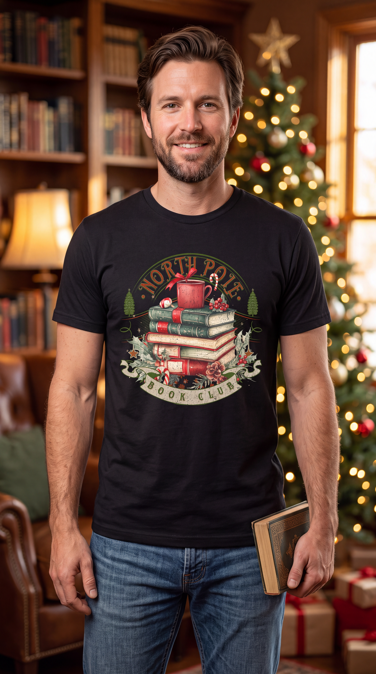 Man wearing black North Pole Book Club Christmas graphic shirt featuring stacked books, holiday mug, candy cane, holly and pinecones.