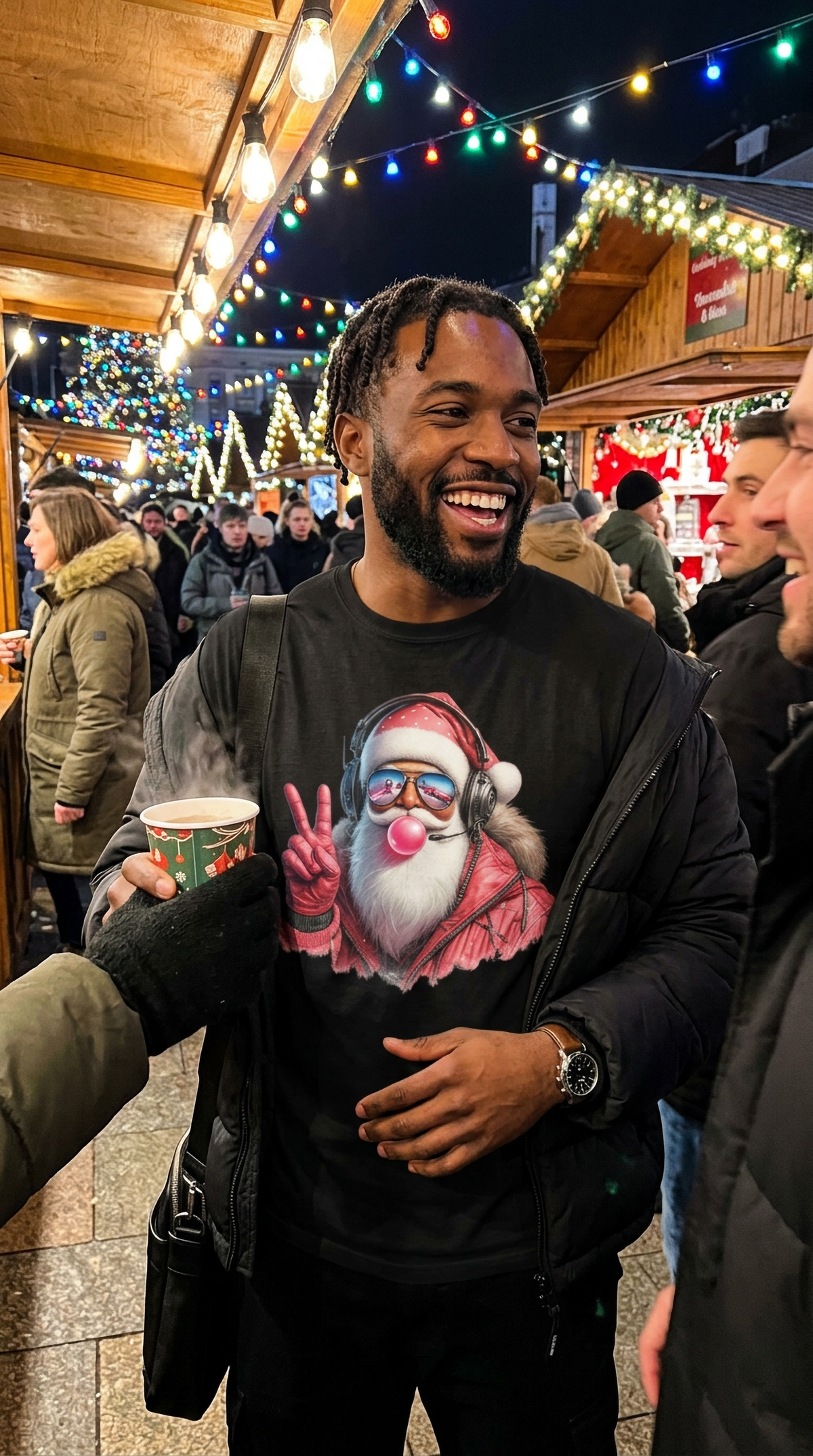 Lifestyle – Black: Man wearing a black Cool Santa Christmas graphic t-shirt at an outdoor nighttime holiday market decorated with colorful Christmas lights.