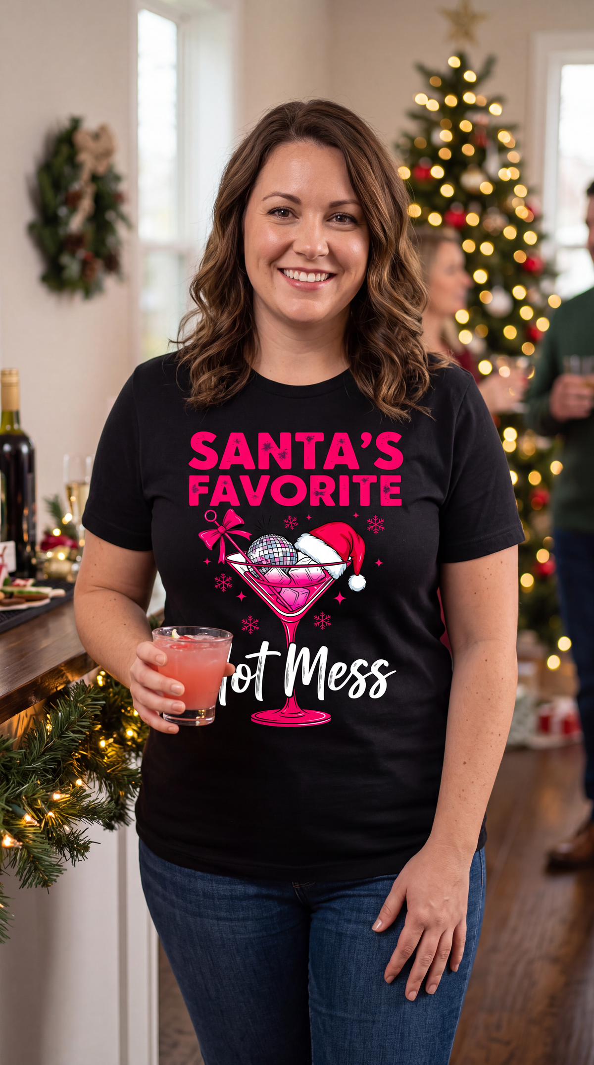 Woman wearing black Santa’s Favorite Hot Mess Christmas shirt near a decorated Christmas tree.