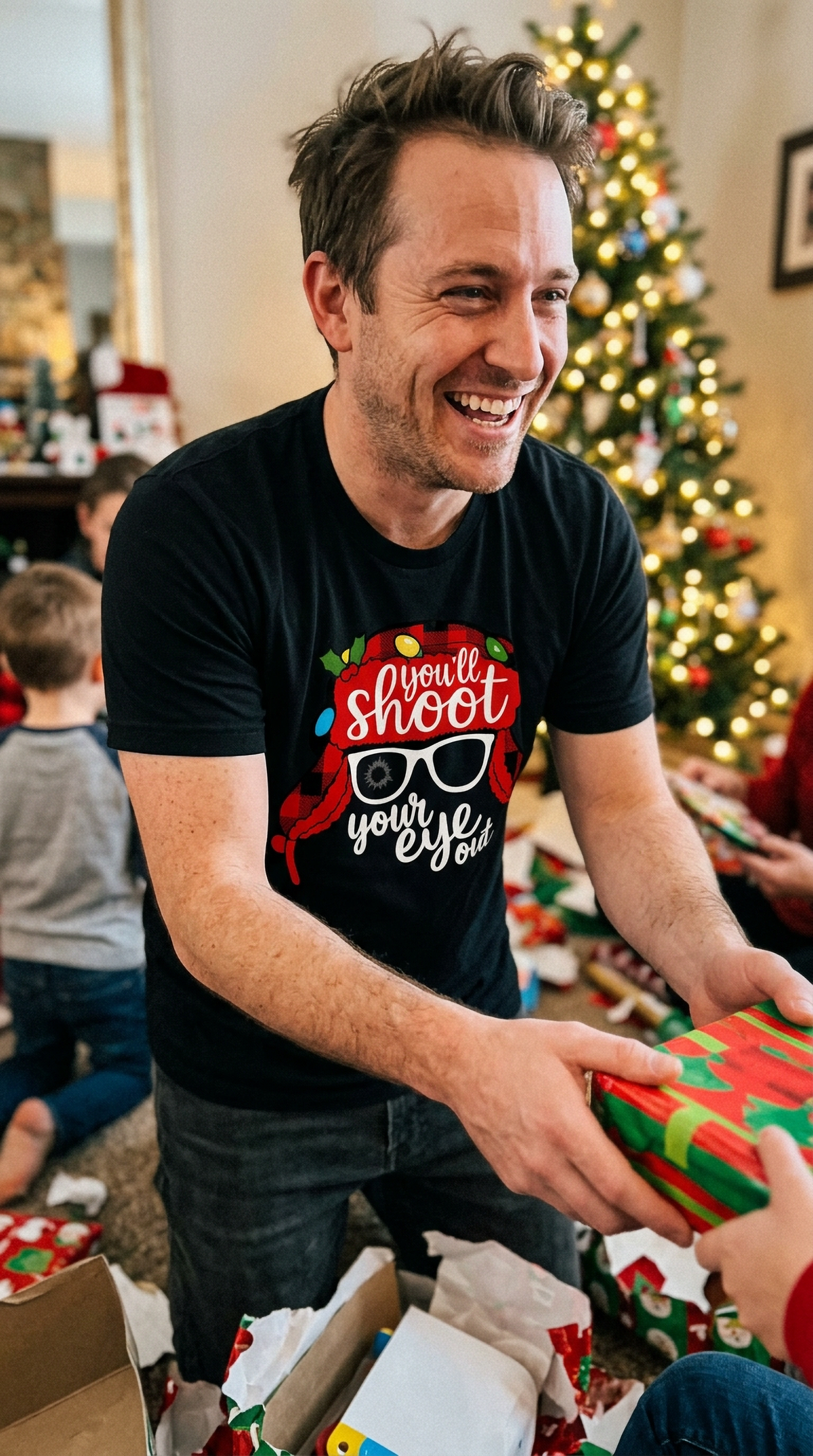 Man wearing black You'll Shoot Your Eye Out Christmas shirt while opening gifts beside a decorated Christmas tree