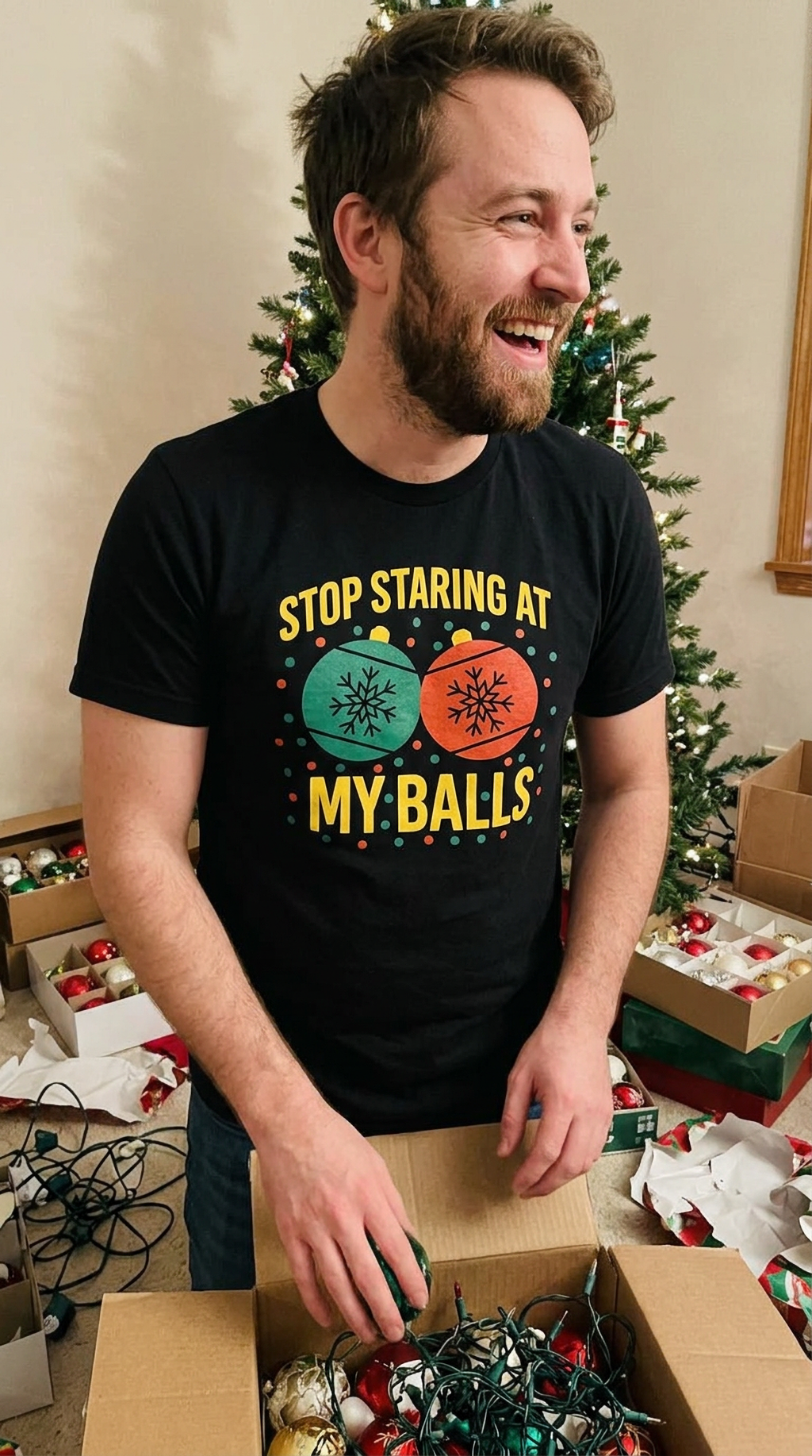 Man wearing black Stop Staring at My Balls funny Christmas t-shirt while decorating for the holidays beside a Christmas tree.