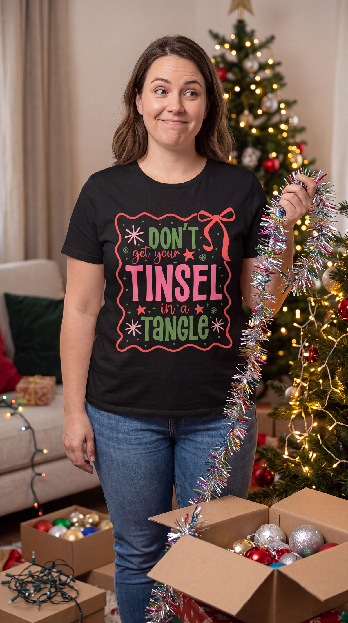 Woman wearing a black Don’t Get Your Tinsel in a Tangle Christmas shirt beside a decorated Christmas tree.