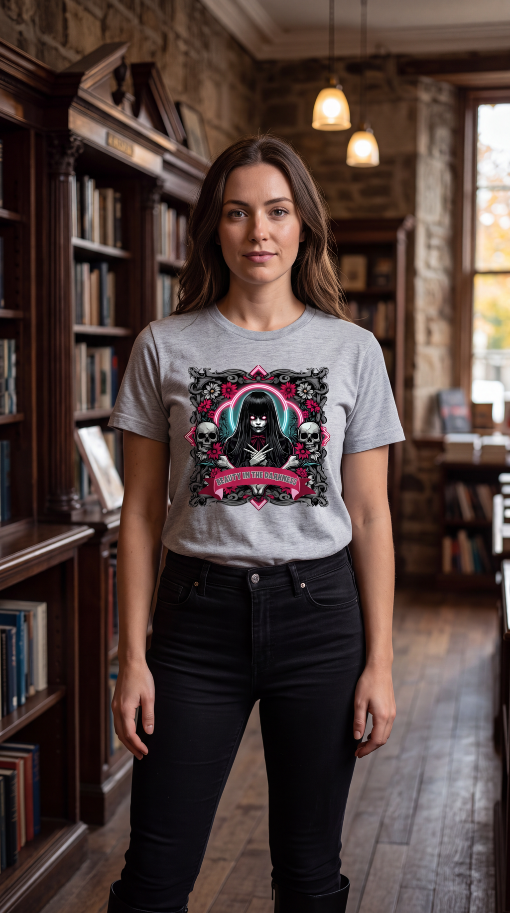 A woman stands in a library wearing a light gray graphic t-shirt with a black, pink, and white gothic design, paired with black jeans.