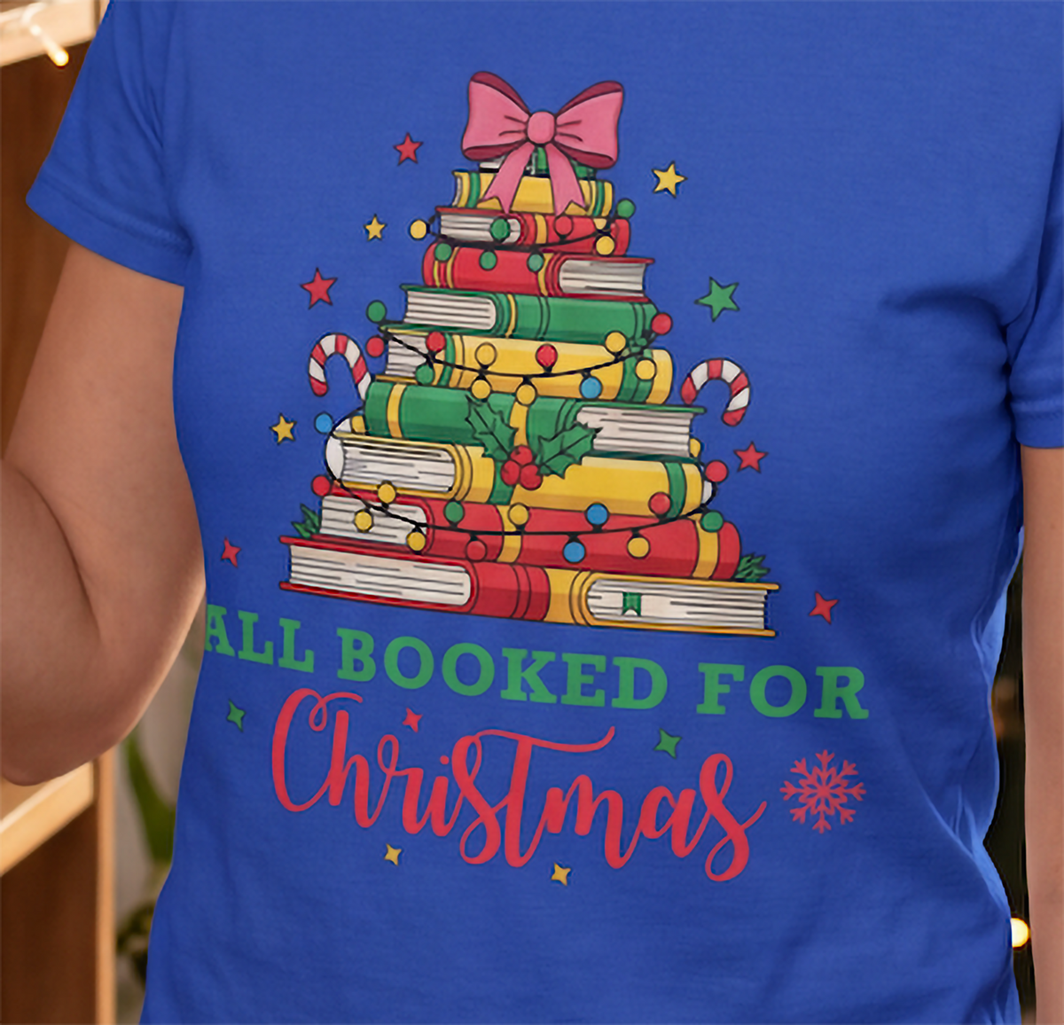 Close-up of blue All Booked for Christmas shirt with stacked books, Christmas lights, candy canes, holly and a bow.