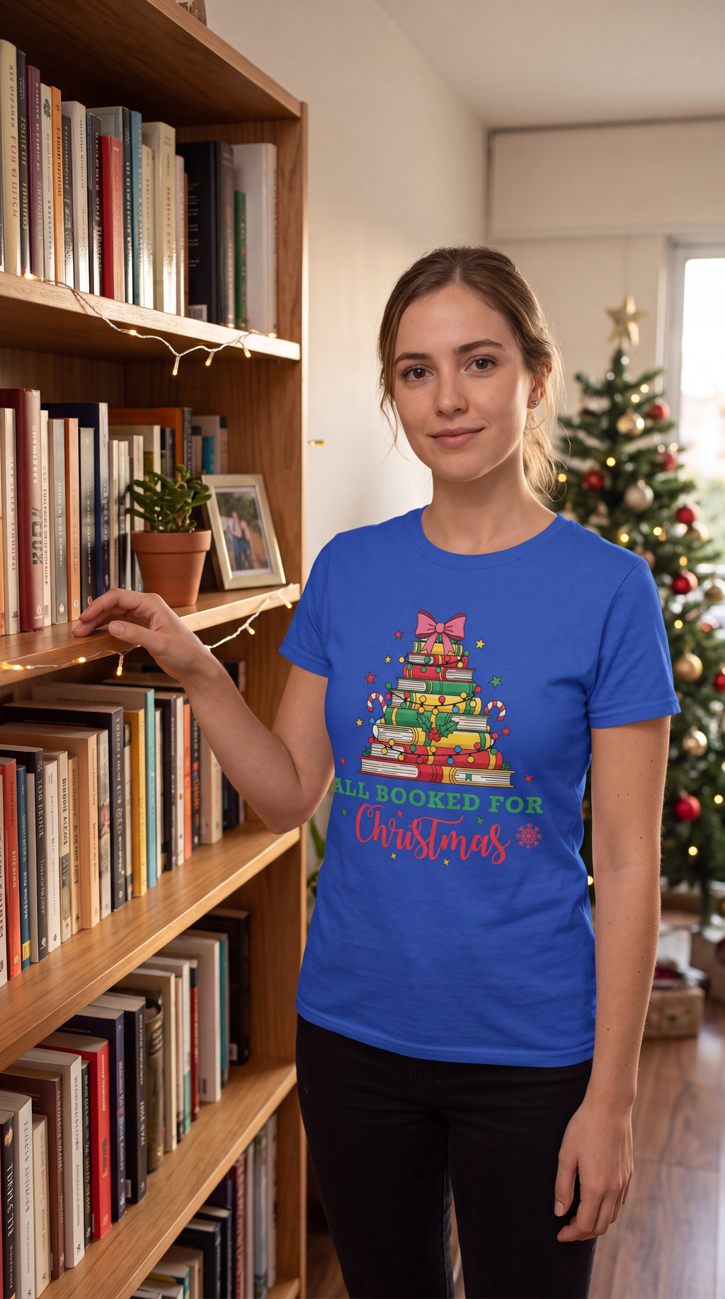 Woman wearing a blue All Booked for Christmas book lover shirt beside bookshelves with a Christmas tree in the background.