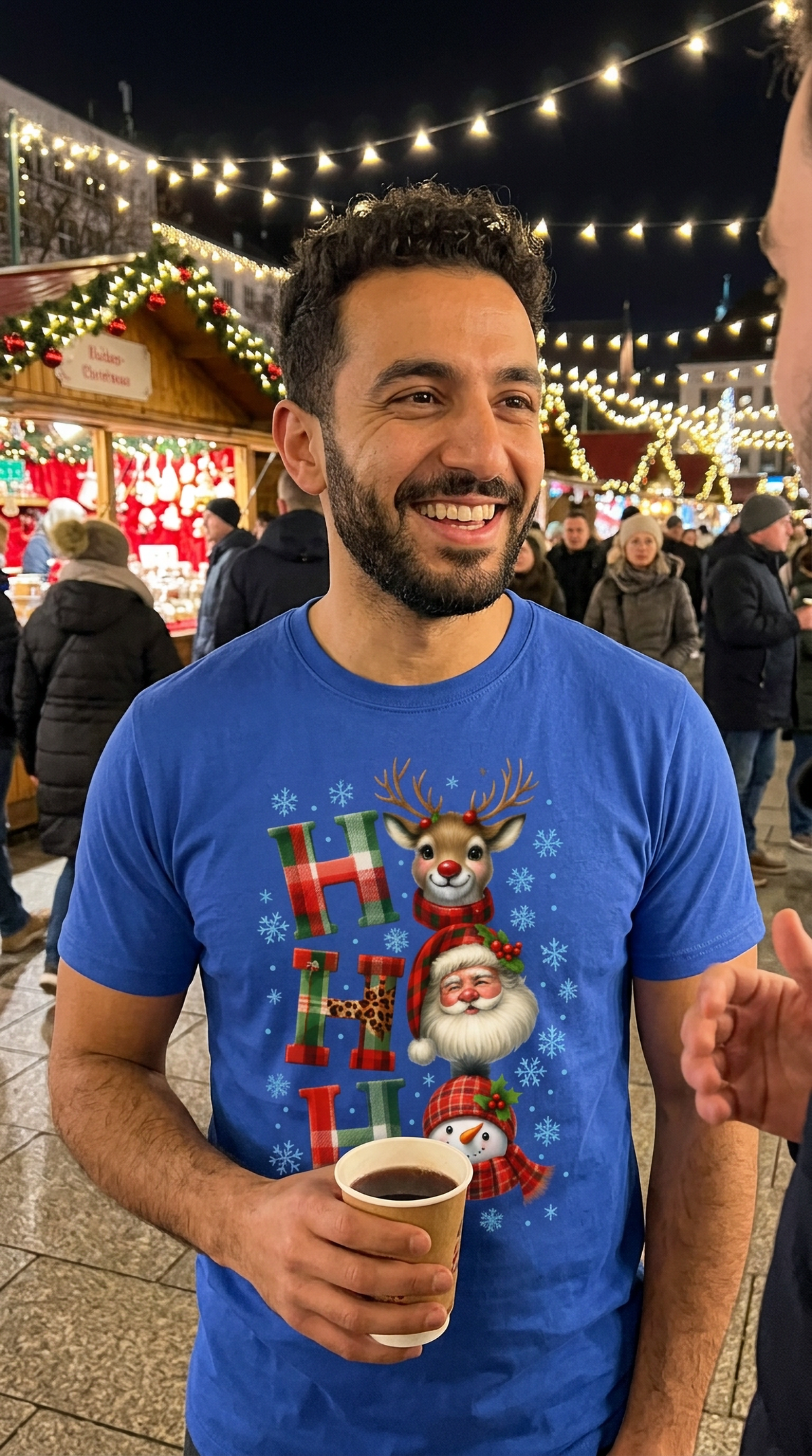 Smiling man wearing blue Ho Ho Ho Christmas shirt while holding a drink at an outdoor holiday gathering.