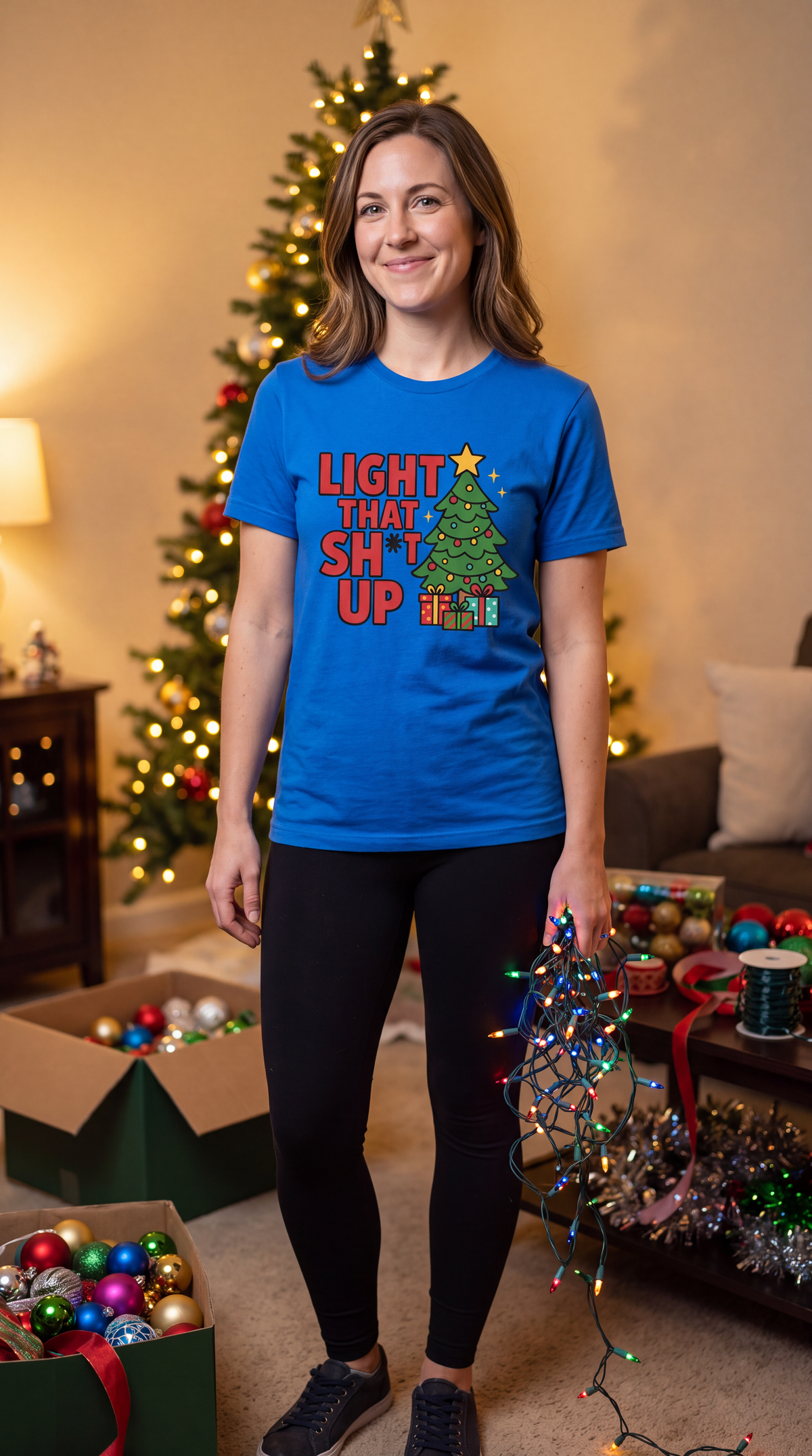 Woman wearing royal blue Light That Shit Up Christmas shirt beside a decorated Christmas tree.