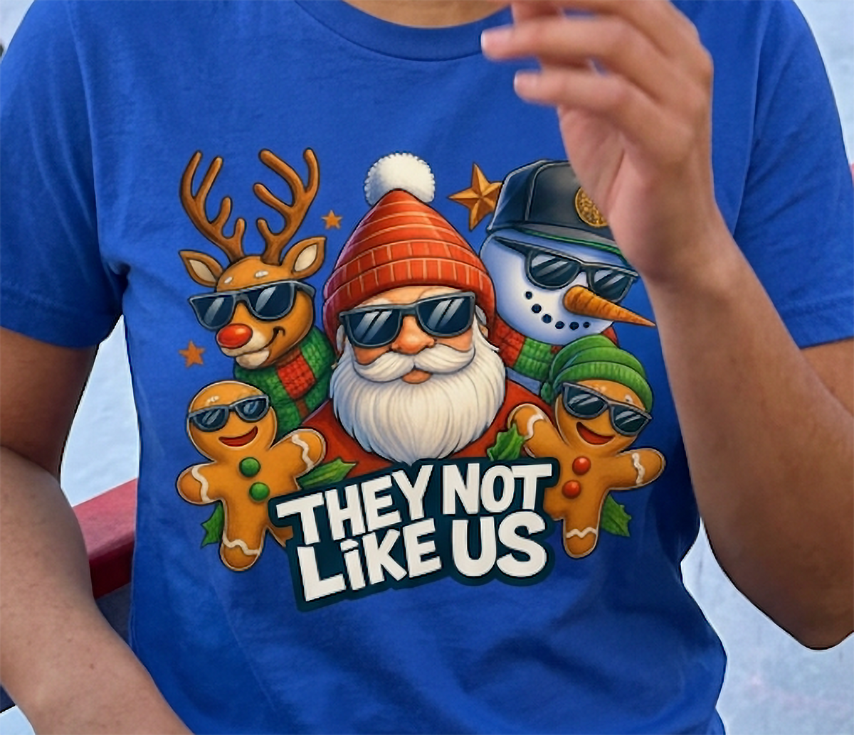 Close-up of blue They Not Like Us Christmas shirt showing funny holiday characters wearing sunglasses and colorful seasonal artwork.