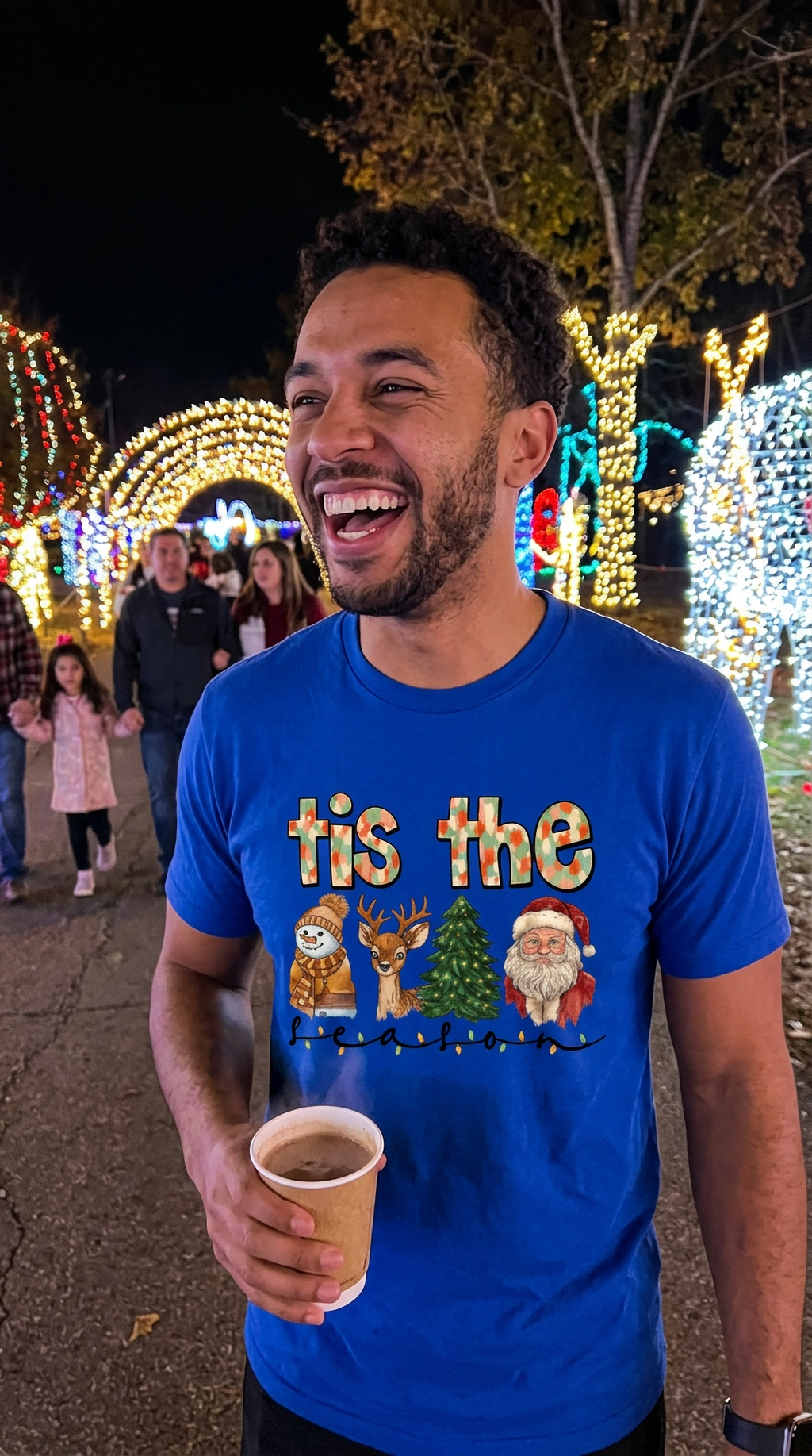 Man wearing blue Tis the Season Christmas graphic t-shirt while holding a drink at an outdoor holiday event illuminated by Christmas lights.