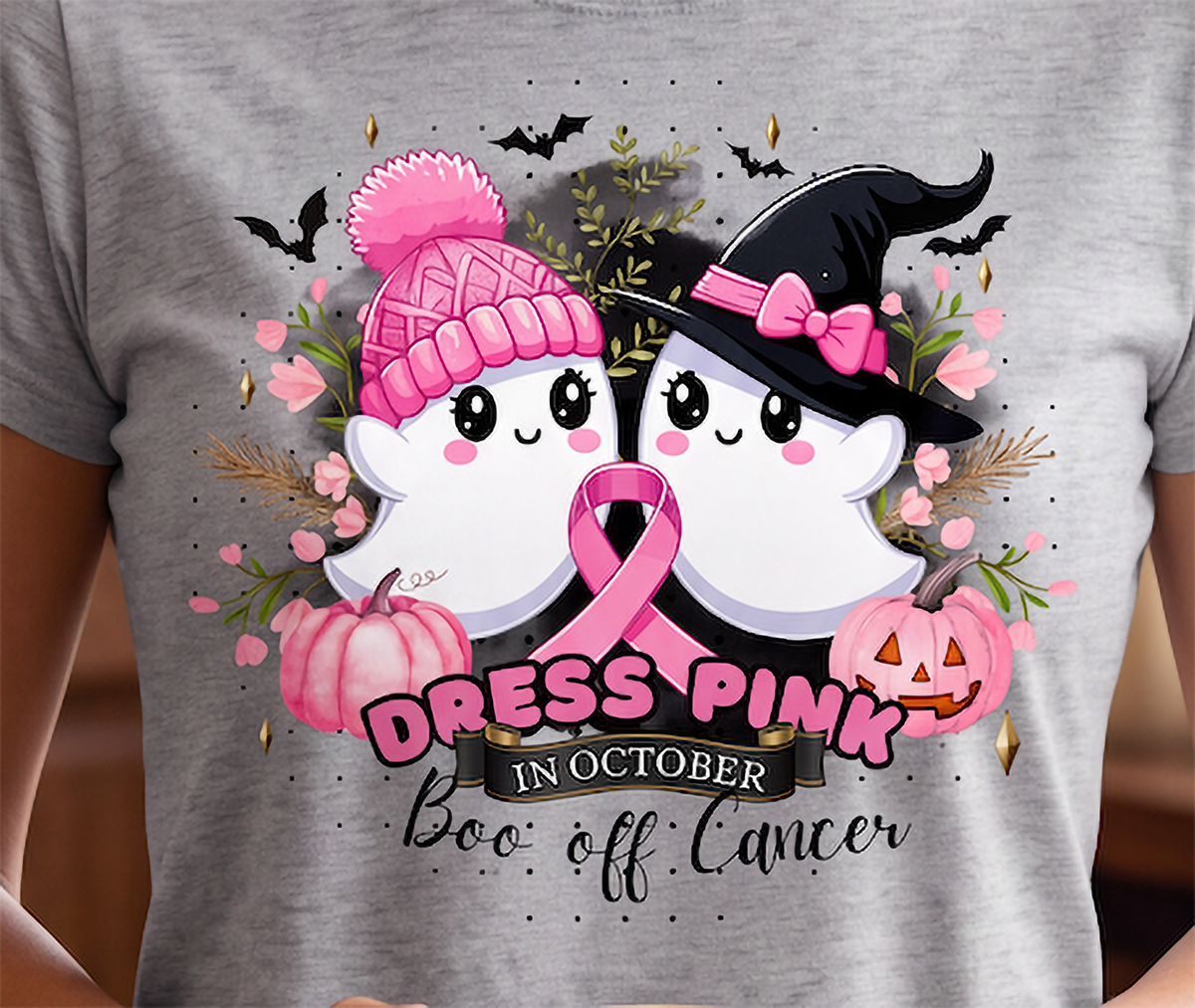 A close-up of a person wearing a heather gray t-shirt with a colorful graphic print. The graphic features two cute cartoon ghosts, one in a pink knit hat and one in a black witch hat, holding a pink ribbon, surrounded by pumpkins, flowers, bats, and the text 'DRESS PINK IN OCTOBER Boo off Cancer'.