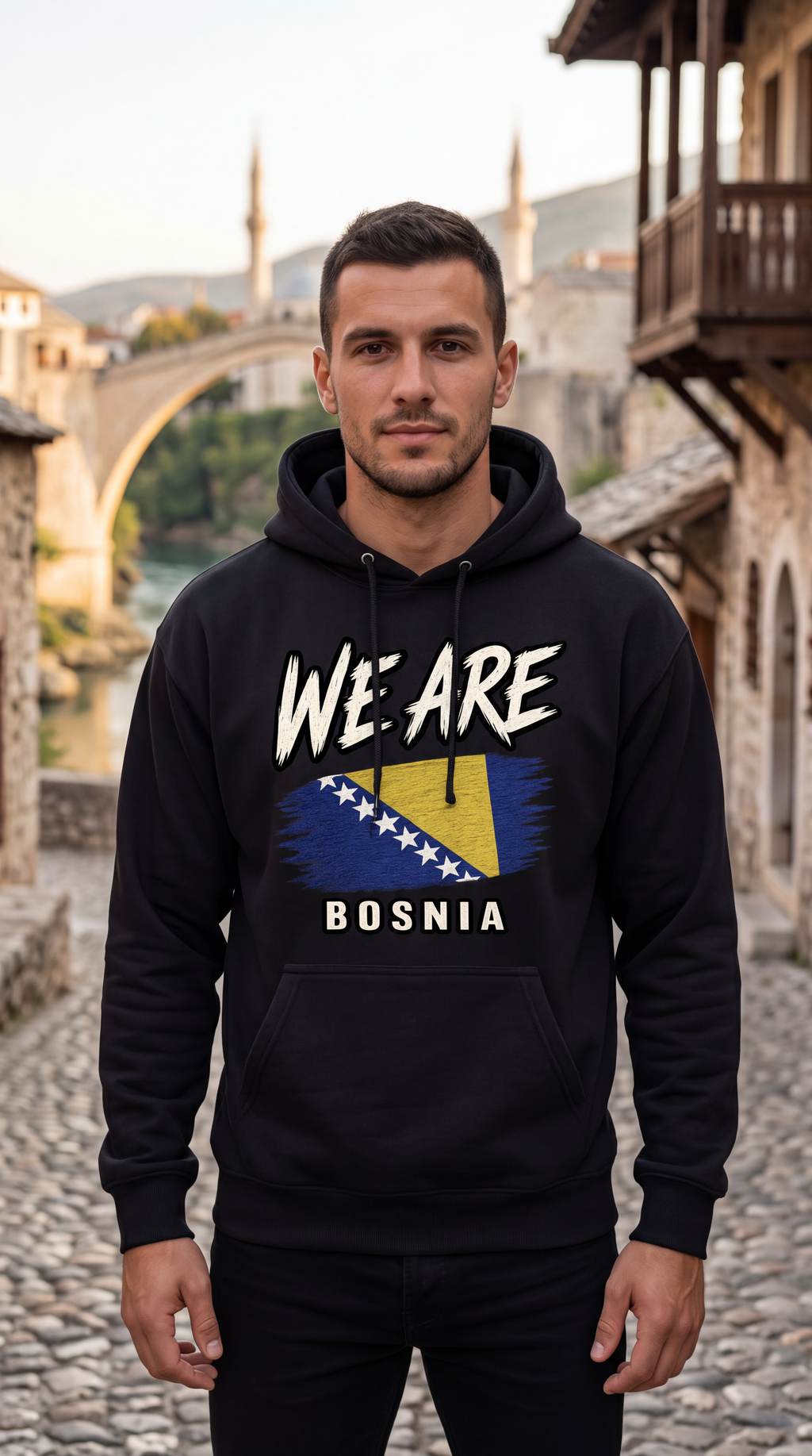 A man wearing a black hoodie with 'WE ARE BOSNIA' and a stylized Bosnia flag graphic on the front, standing on a cobblestone street with a stone bridge and buildings in the background.
