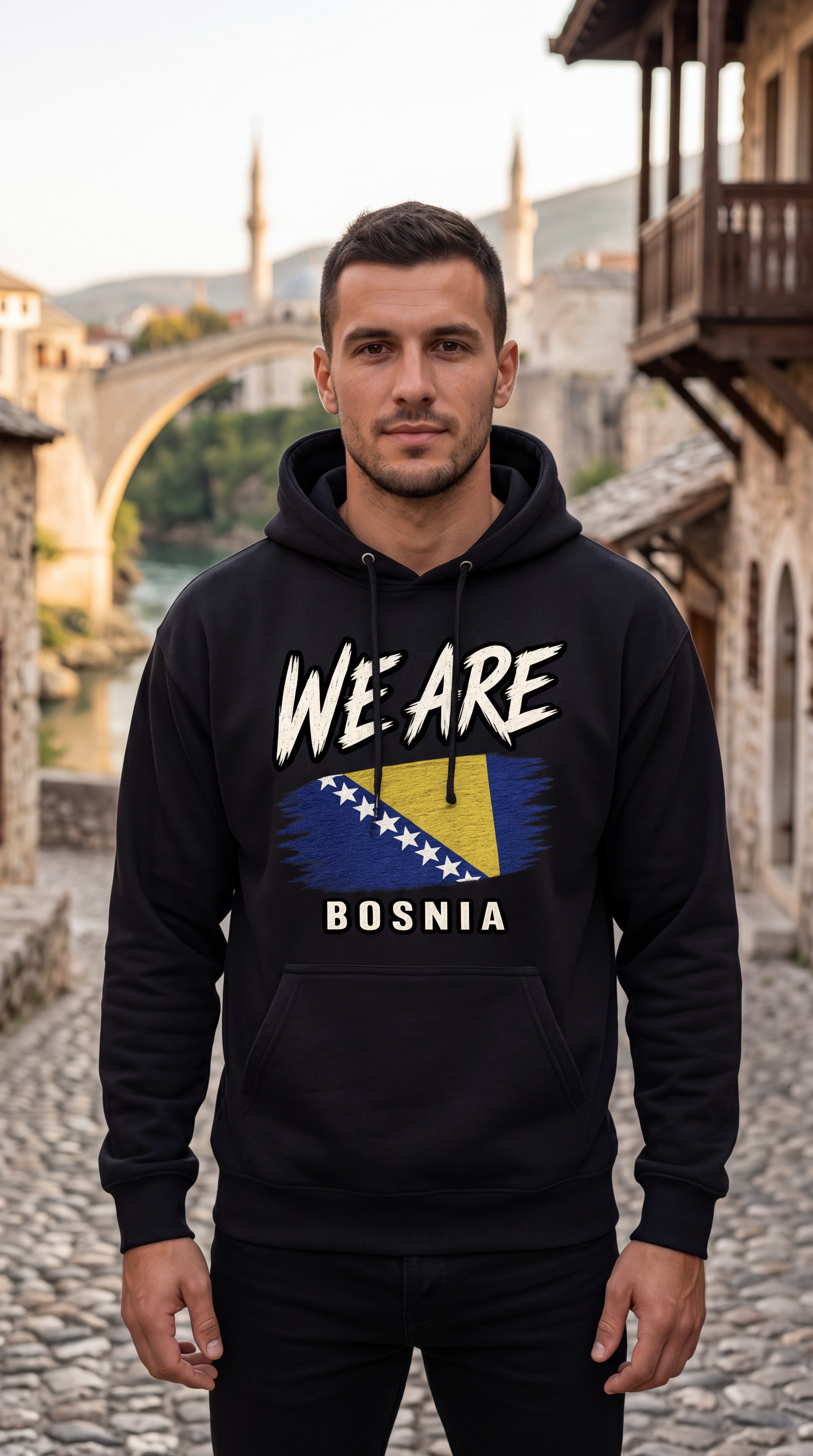 A man wearing a black hoodie with 'WE ARE BOSNIA' and a stylized Bosnia flag graphic on the front, standing on a cobblestone street with a stone bridge and buildings in the background.