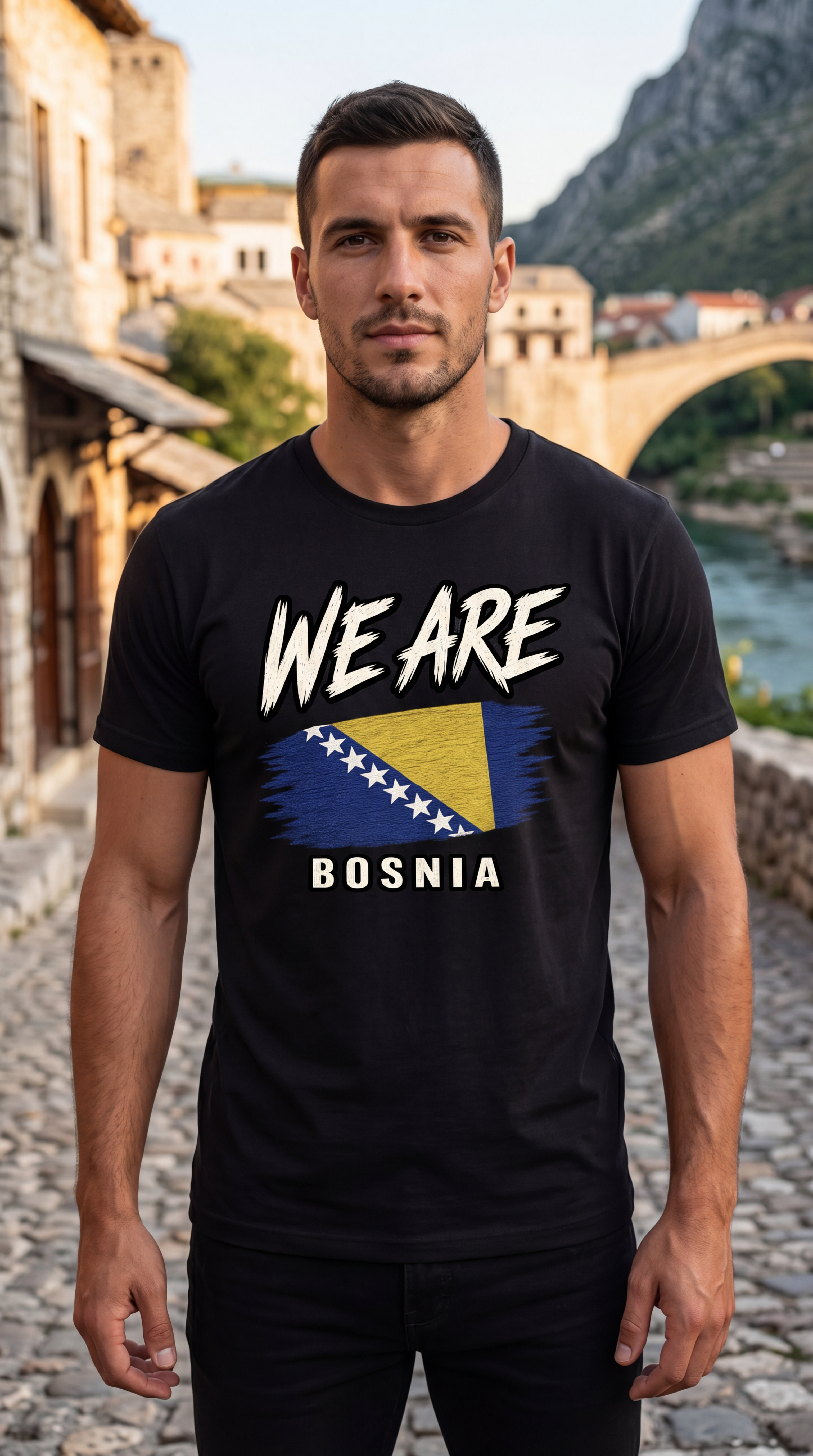 A man wearing a black graphic t-shirt with 'WE ARE BOSNIA' and the Bosnia flag design, standing outdoors in front of a scenic stone bridge and river background.