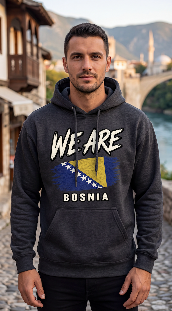 A man wearing a dark gray hoodie with a graphic print that reads 'WE ARE BOSNIA' and features the Bosnia flag, standing outdoors in front of a scenic background with a bridge and river.