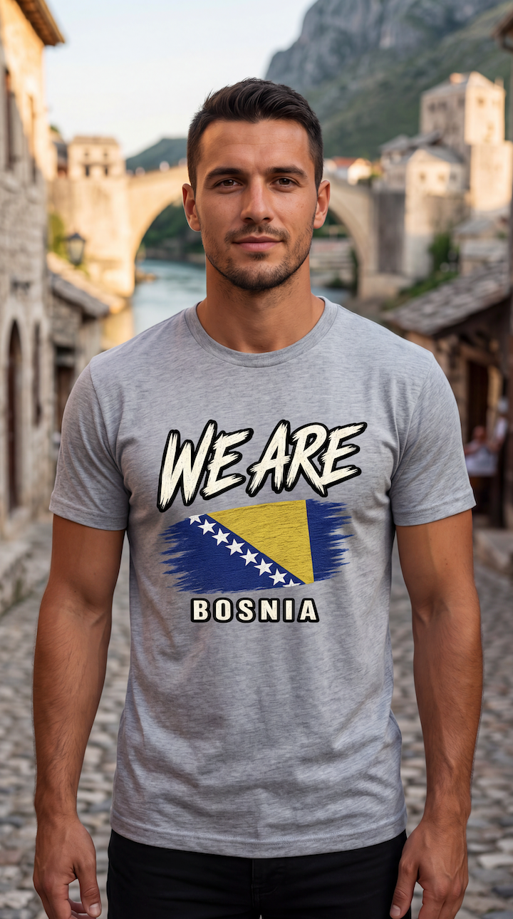 A man wearing a heather gray t-shirt with a graphic that reads 'WE ARE BOSNIA' and features the Bosnia flag, standing in front of a scenic stone bridge and mountain background.