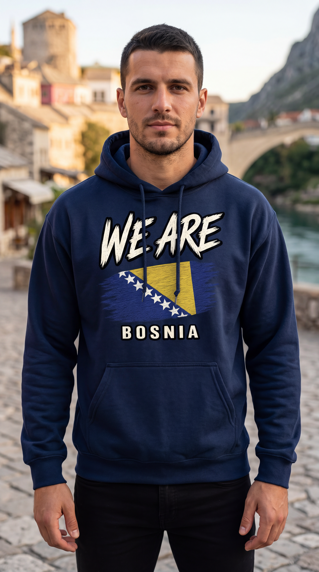 A man wearing a navy blue hoodie with a graphic print that reads 'WE ARE BOSNIA' and features the Bosnia flag, standing outdoors in front of a scenic background of a bridge and historic buildings.