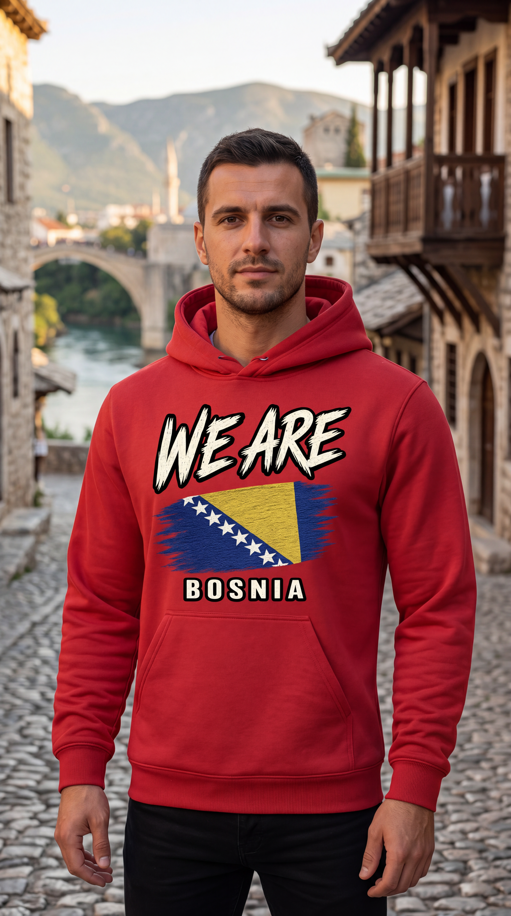 A man wearing a red hoodie with a graphic print that reads 'WE ARE BOSNIA' and features the Bosnia flag, standing on a cobblestone street in front of traditional buildings and a river.