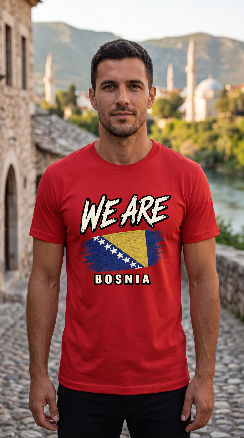 A man wearing a red graphic t-shirt with 'WE ARE BOSNIA' and the Bosnia flag printed on the front, standing outdoors on a cobblestone street with a scenic background.