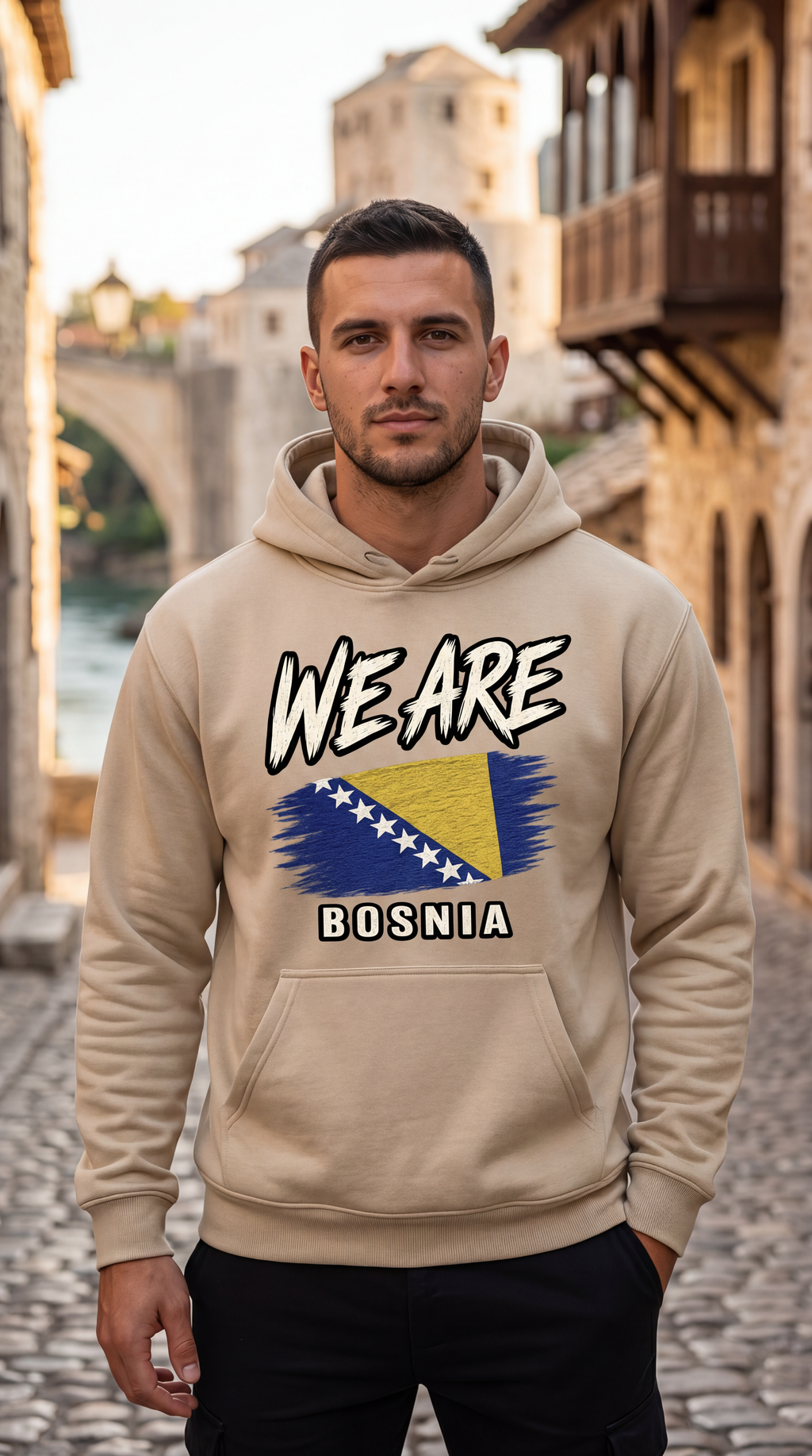 A man wearing a beige hoodie with a graphic print of the Bosnia flag and the text 'WE ARE BOSNIA' stands on a cobblestone street with a blurred background of historic buildings and a bridge.