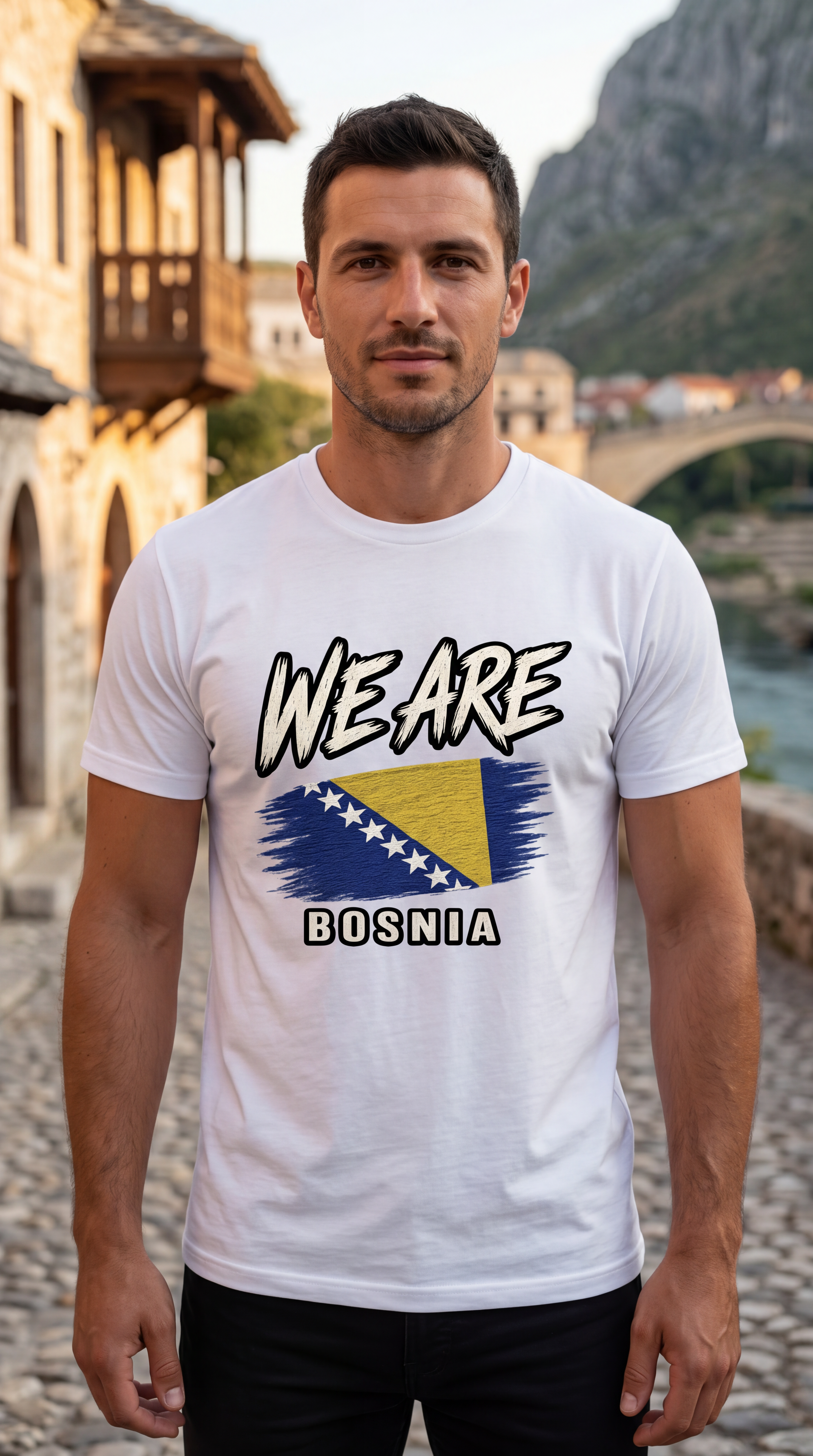 A man wearing a white graphic t-shirt with 'WE ARE BOSNIA' and the Bosnia flag printed on the front, standing outdoors in front of a scenic stone bridge and mountain backdrop.