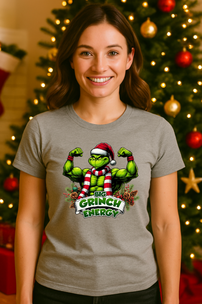 Woman wearing a gray holiday muscle-energy graphic t-shirt, smiling in front of a decorated Christmas tree with warm festive lights and ornaments.