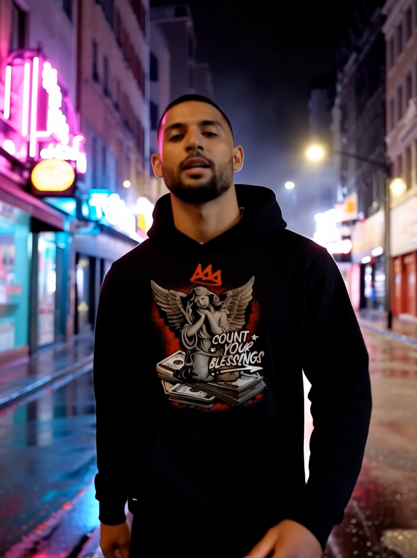Man wearing black streetwear hoodie with angel and Count Your Blessings graphic in city street at night