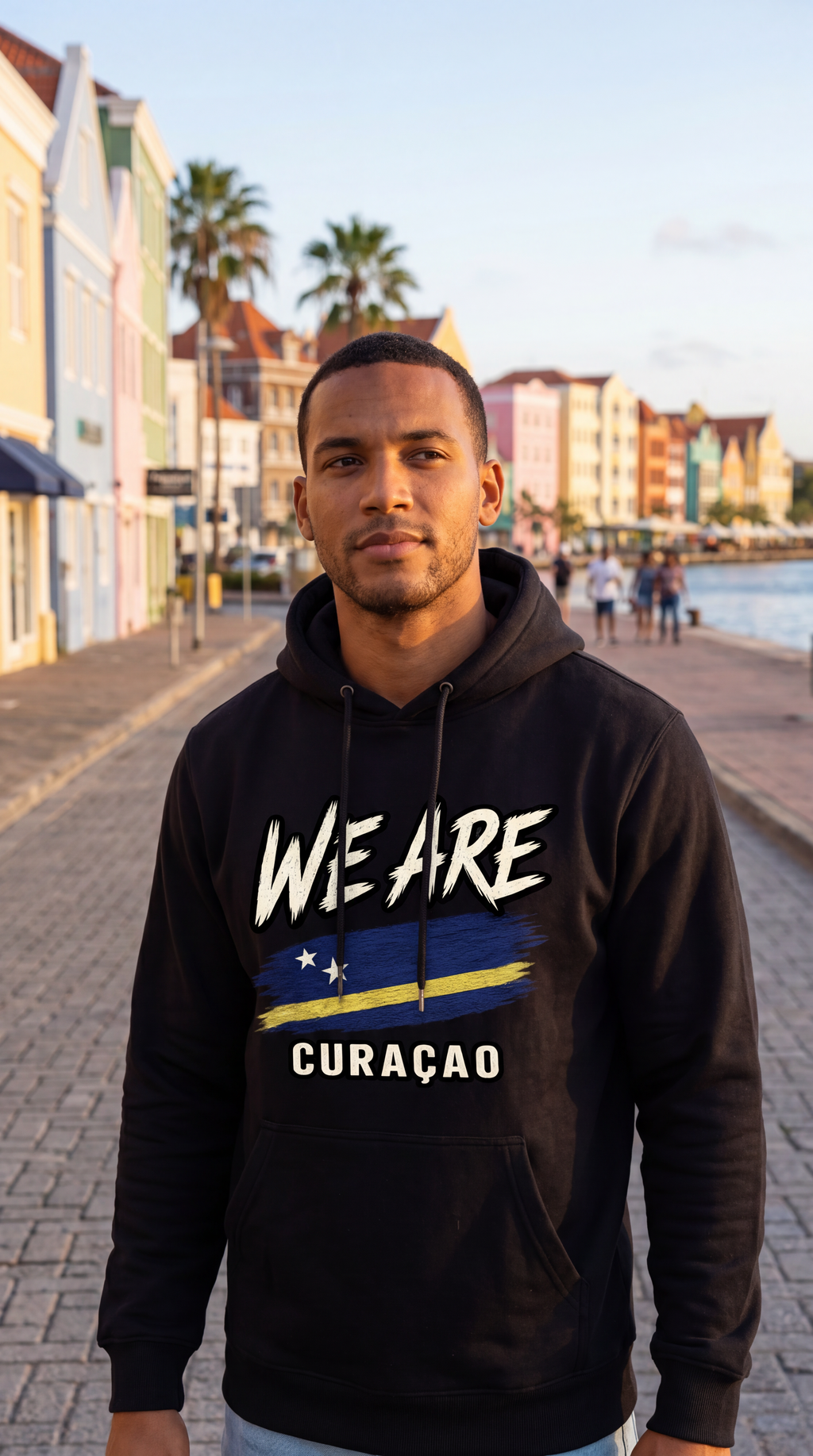 A man wearing a black hoodie with 'WE ARE CURAÇAO' and a yellow-blue flag graphic, standing on a waterfront promenade with colorful buildings in the background.