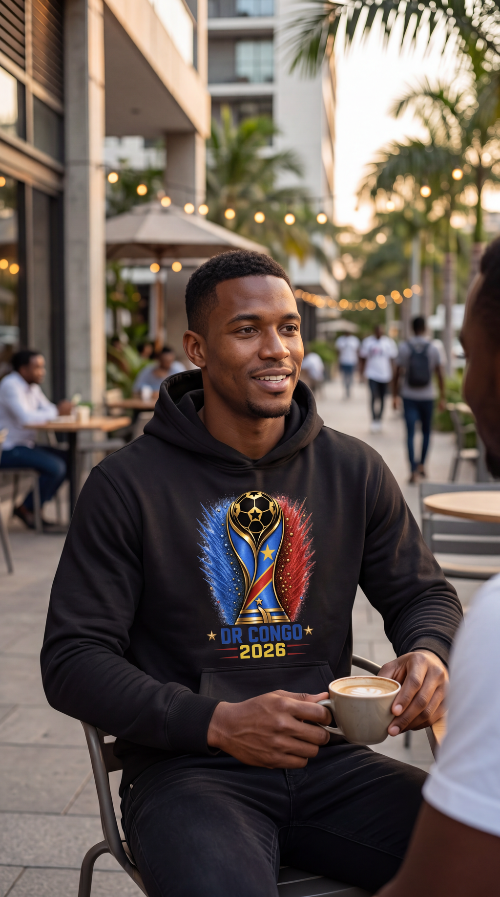 Man wearing a black hoodie with a colorful design, sitting outdoors with people and buildings in the background.