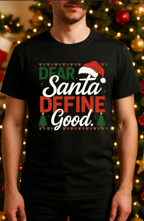 Person wearing a black “Dear Santa Define Good” Christmas graphic T-shirt standing in front of a decorated Christmas tree with warm holiday lights.