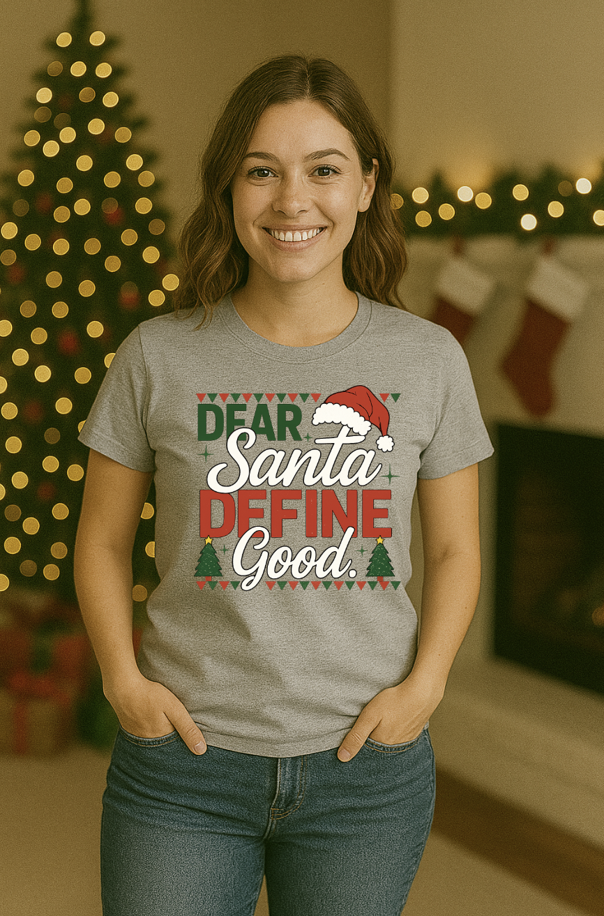 Woman wearing a gray “Dear Santa Define Good” Christmas T-shirt, standing in front of a decorated Christmas tree and festive holiday stockings.