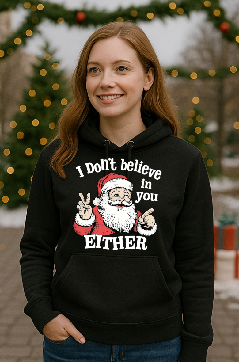Woman wearing a black Christmas hoodie with a sarcastic Santa Claus graphic and the phrase “I Don’t Believe in You Either,” standing outdoors with decorated Christmas trees in the background.