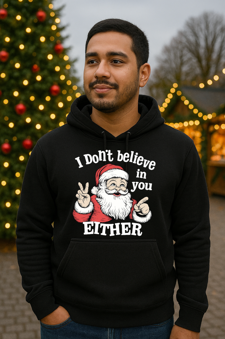 Man wearing a black Christmas hoodie with a sarcastic Santa Claus graphic and the phrase “I Don’t Believe in You Either,” standing outdoors in front of a decorated Christmas tree with lights.