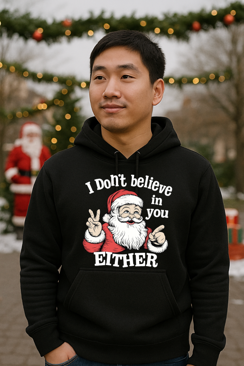 Man wearing a black Christmas hoodie with a sarcastic Santa Claus graphic and the phrase “I Don’t Believe in You Either,” standing outdoors with festive Christmas decorations in the background.