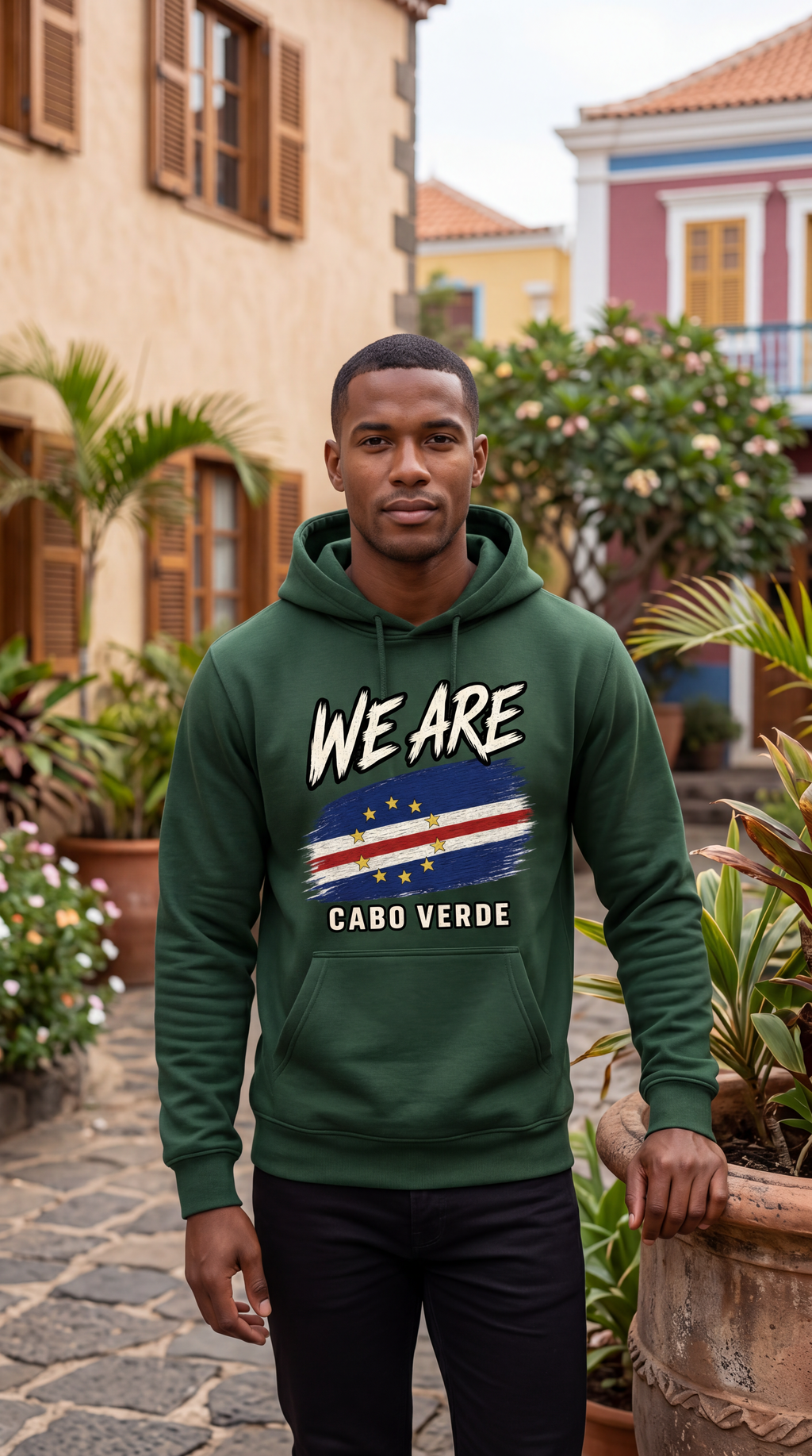 A man wearing a dark green hoodie with a graphic print that reads 'WE ARE CABO VERDE' and features the Cabo Verde flag, standing in an outdoor courtyard with plants and buildings in the background.