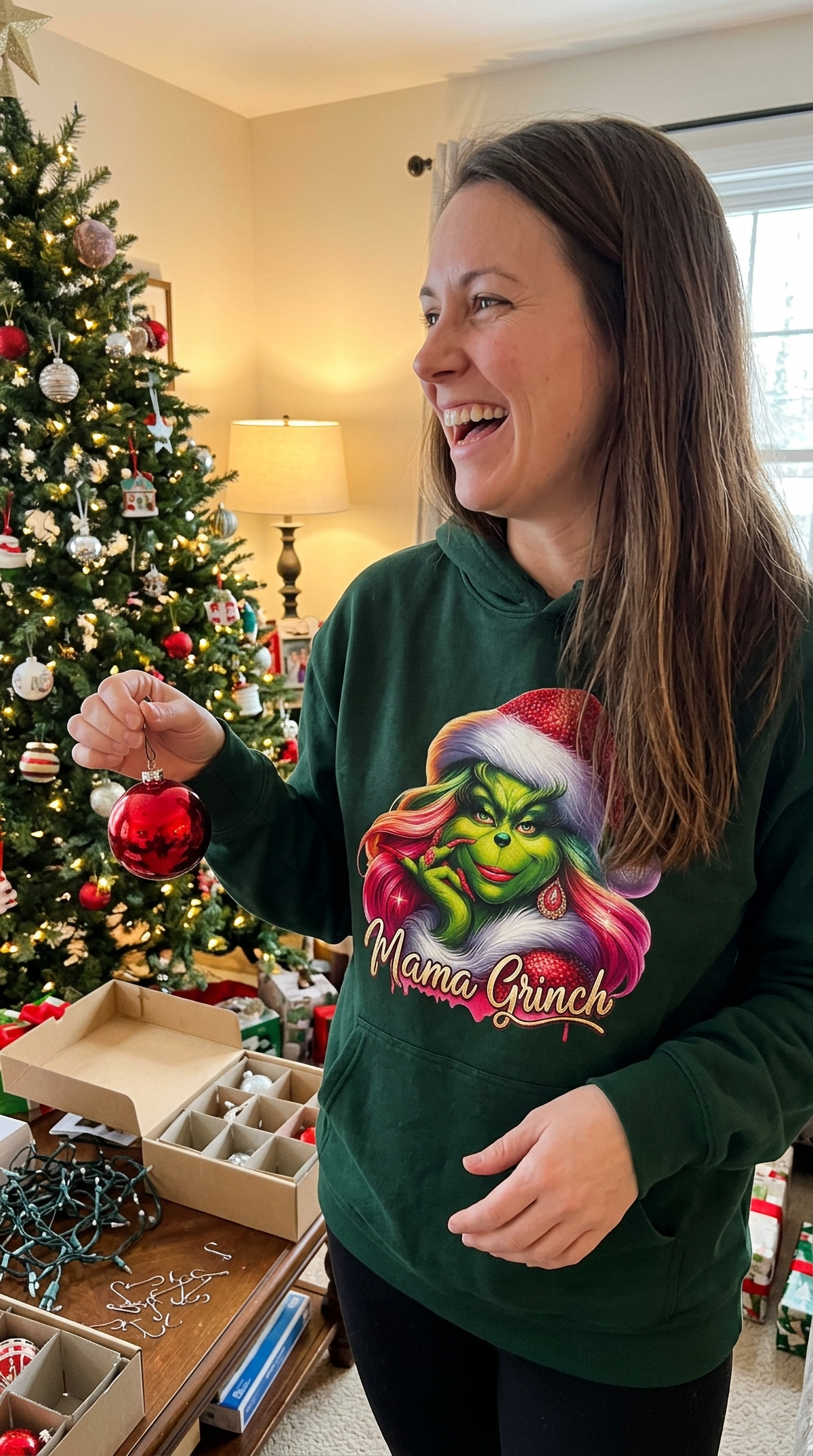 Woman wearing forest green Mama Grinch hoodie beside a decorated Christmas tree