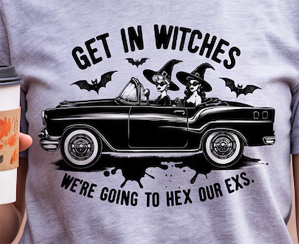 A close-up of a person wearing a heather gray t-shirt with a black graphic print of a vintage car driven by two witches, with the text 'GET IN WITCHES WE'RE GOING TO HEX OUR EXS.' A hand holding a coffee cup is visible on the left side.