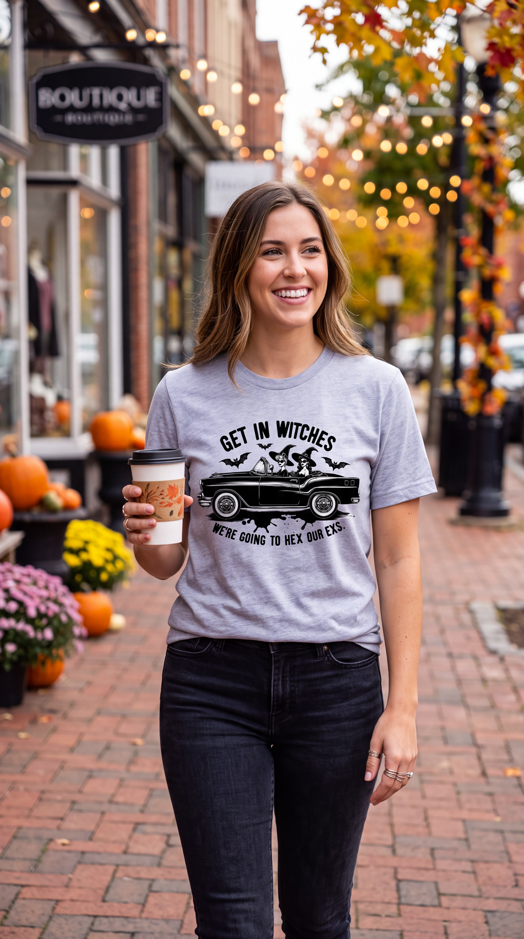 A woman stands on a brick sidewalk wearing a light gray graphic t-shirt with a black witch car design and the text 'GET IN WITCHES WE'RE GOING TO HEX OUR EXS'. She is holding a coffee cup in an outdoor autumn street setting with pumpkins and fall decorations.