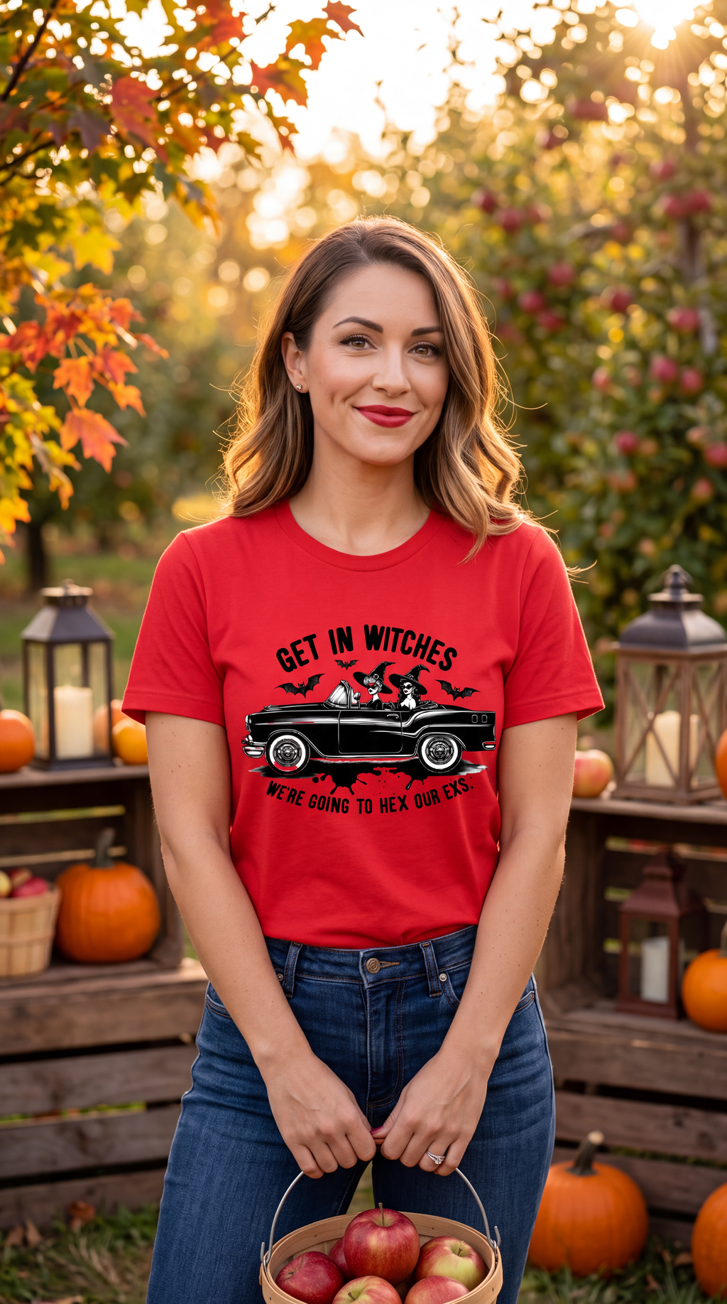 A woman stands outdoors in a fall garden setting wearing a bright red graphic T-shirt and blue jeans, holding a basket of apples. The shirt features a black car with bats and the text 'GET IN WITCHES WE'RE GOING TO HEX OUR EXS'.