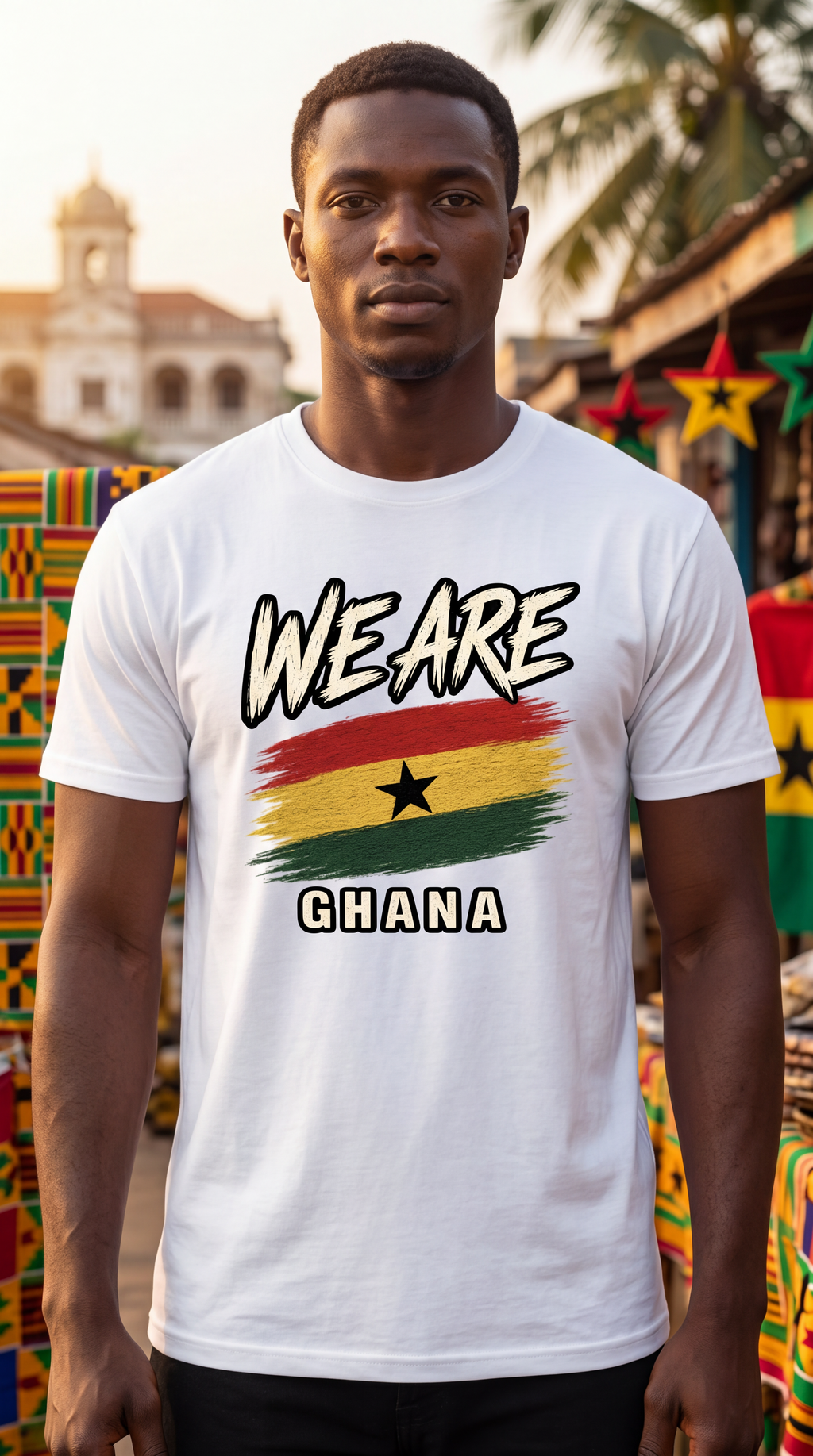 A man wearing a white graphic t-shirt with 'WE ARE GHANA' and the Ghana flag design, standing in front of a blurred outdoor market background.