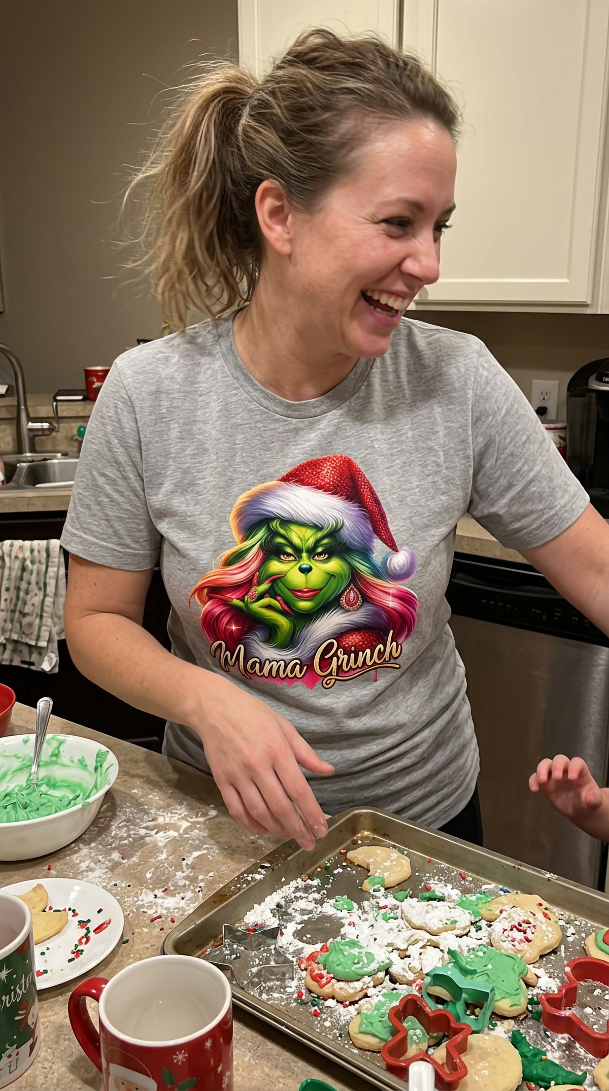 Woman wearing heather gray Mama Grinch Christmas shirt while wrapping presents at a festive holiday gathering.