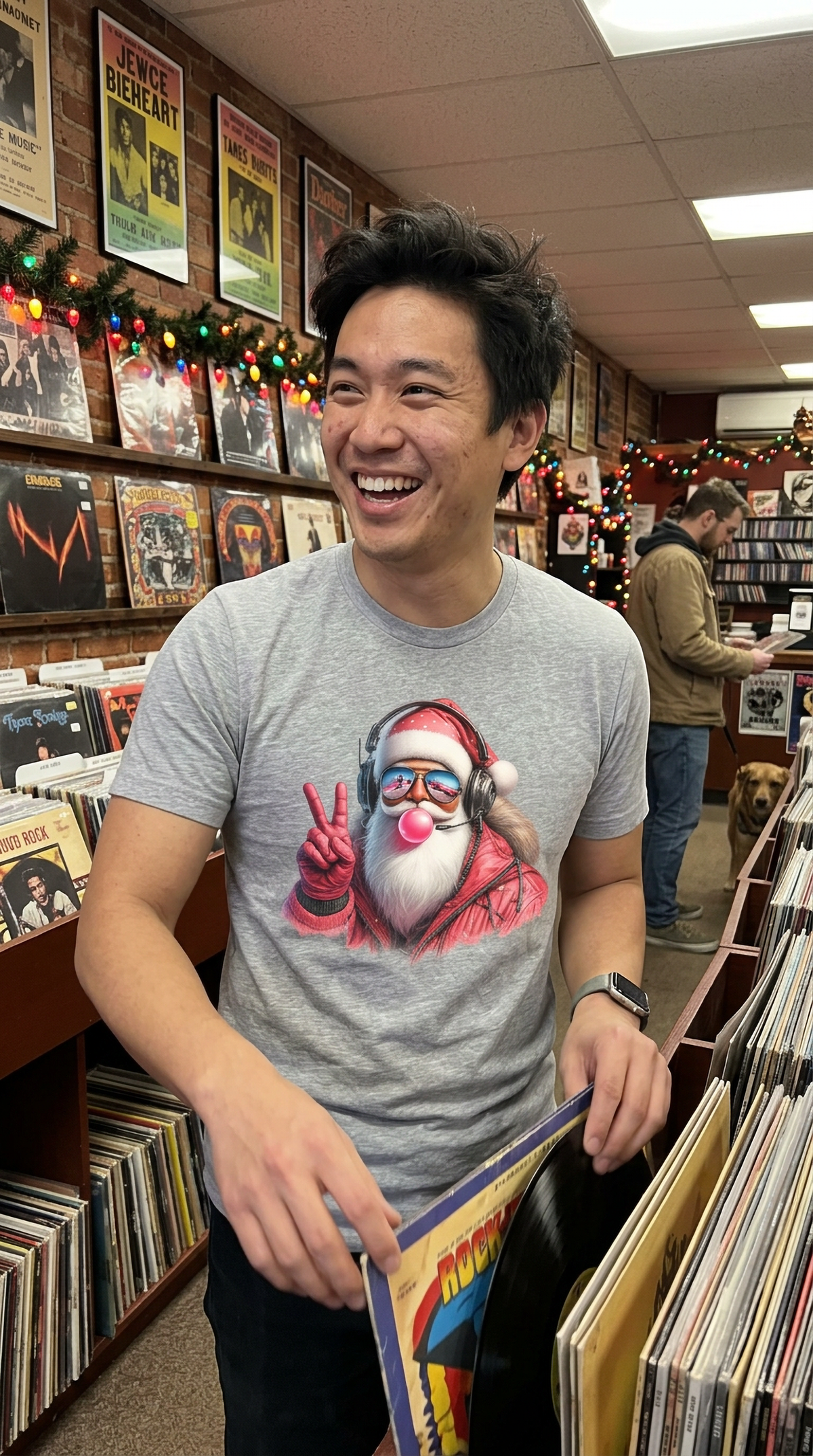 Lifestyle – Heather Gray: Smiling man wearing a heather gray Cool Santa Christmas graphic t-shirt while browsing records inside a festive music store decorated with Christmas lights.