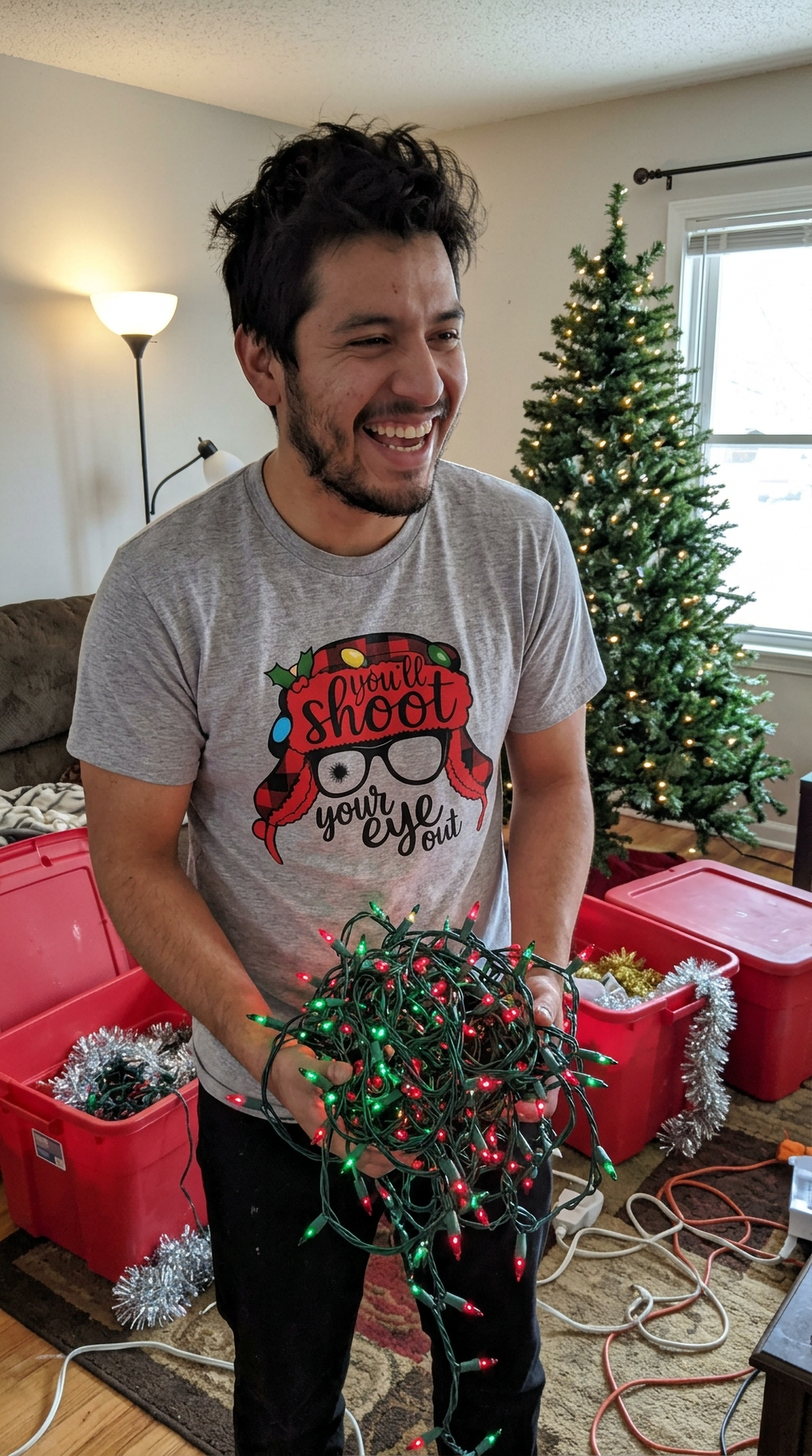 Man wearing heather gray funny Christmas movie quote shirt while celebrating beside a Christmas tree