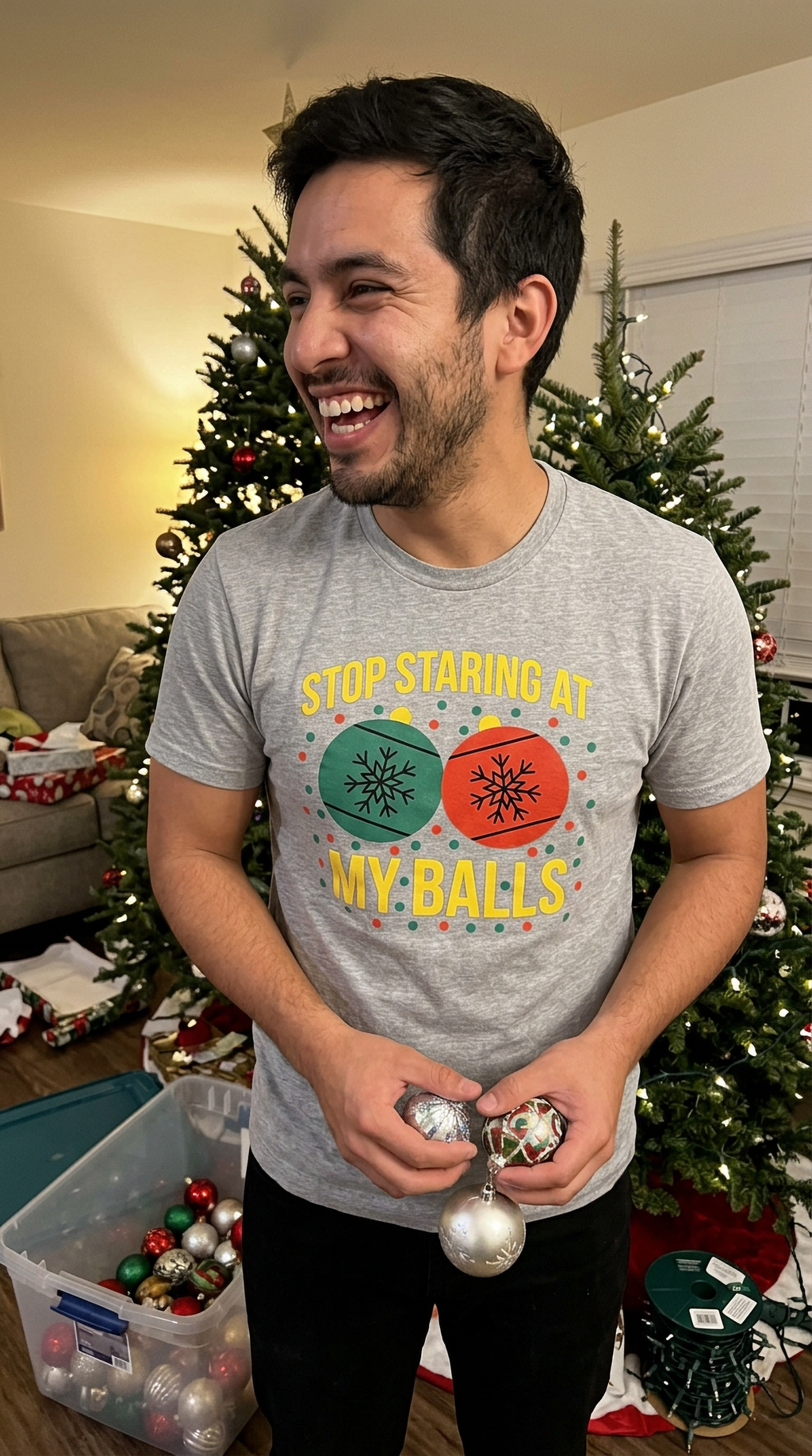 Man wearing heather gray funny Christmas ornament shirt while standing near a decorated Christmas tree.