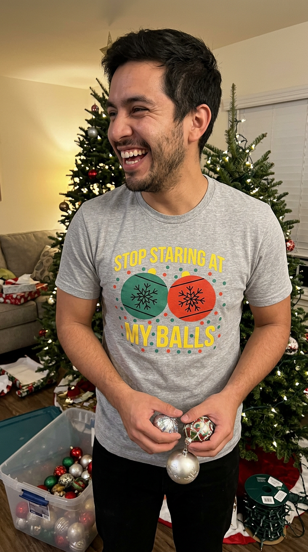 Man wearing heather gray funny Christmas ornament shirt while standing near a decorated Christmas tree.