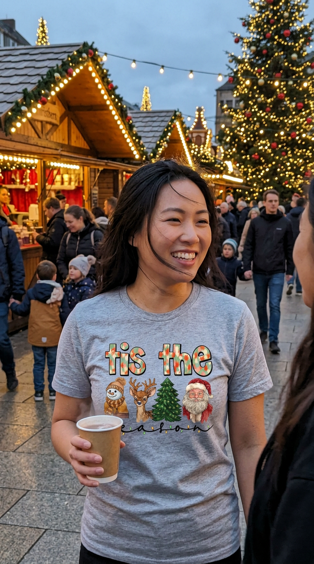 Woman wearing heather gray Tis the Season Christmas graphic t-shirt while enjoying a holiday market with festive decorations and Christmas trees.